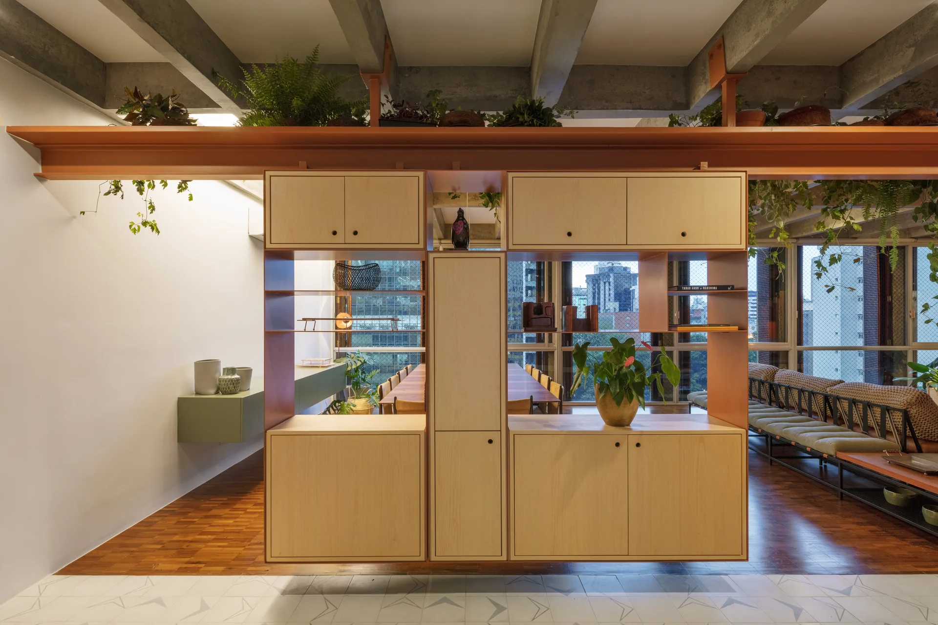 A modern open-concept room features a central light wood cabinet divider beneath an orange steel beam and exposed concrete ceiling beams.