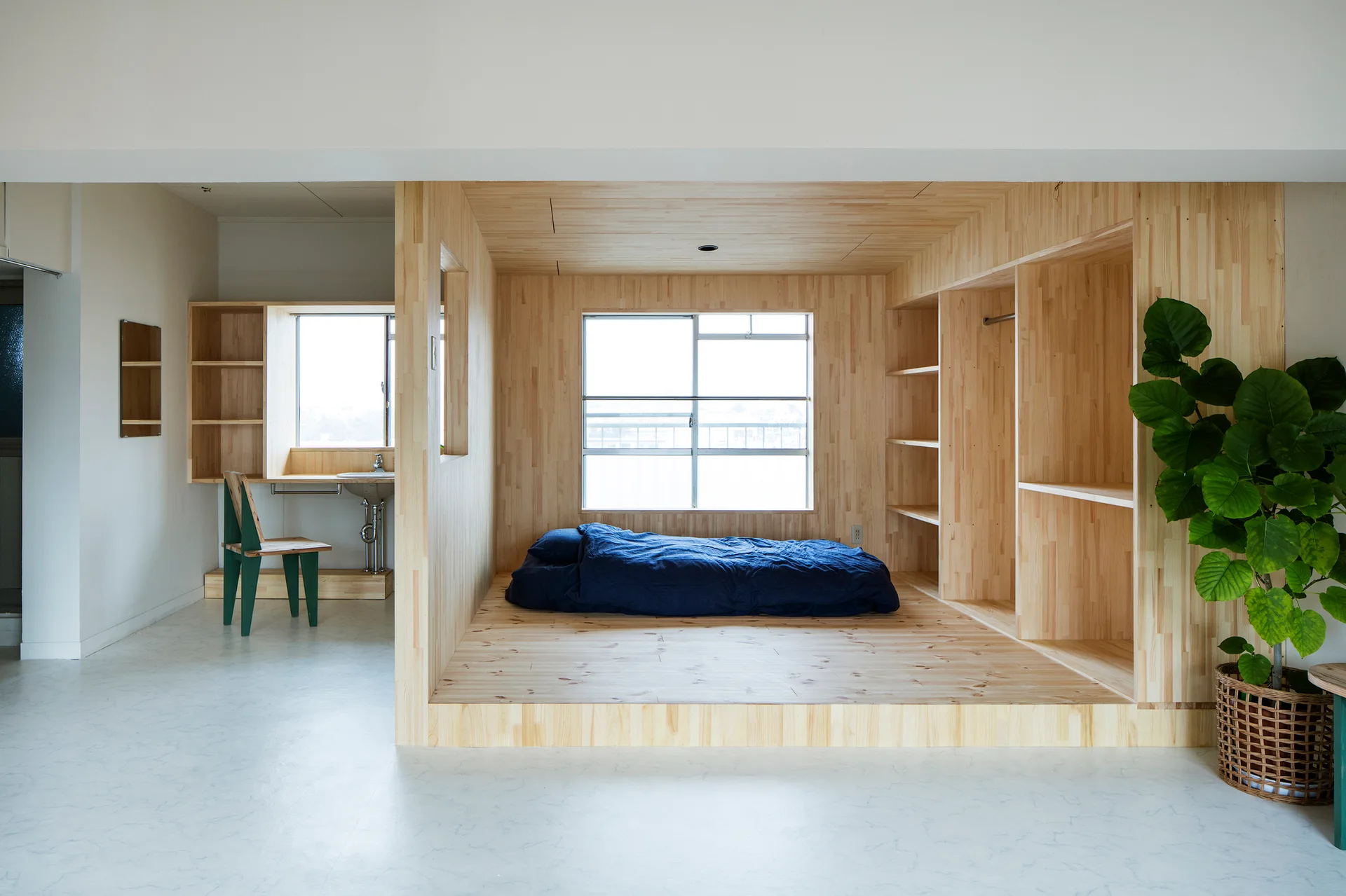 A light wood paneled bedroom alcove features a navy blue mattress on a raised platform next to built-in shelves and a window.