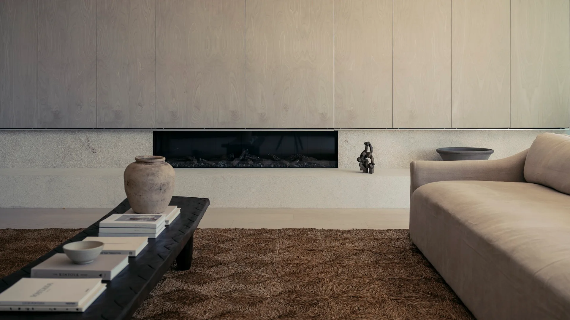 A minimalist living room featuring a recessed linear fireplace below light wood wall panels, a low beige sofa, and a textured brown rug.
