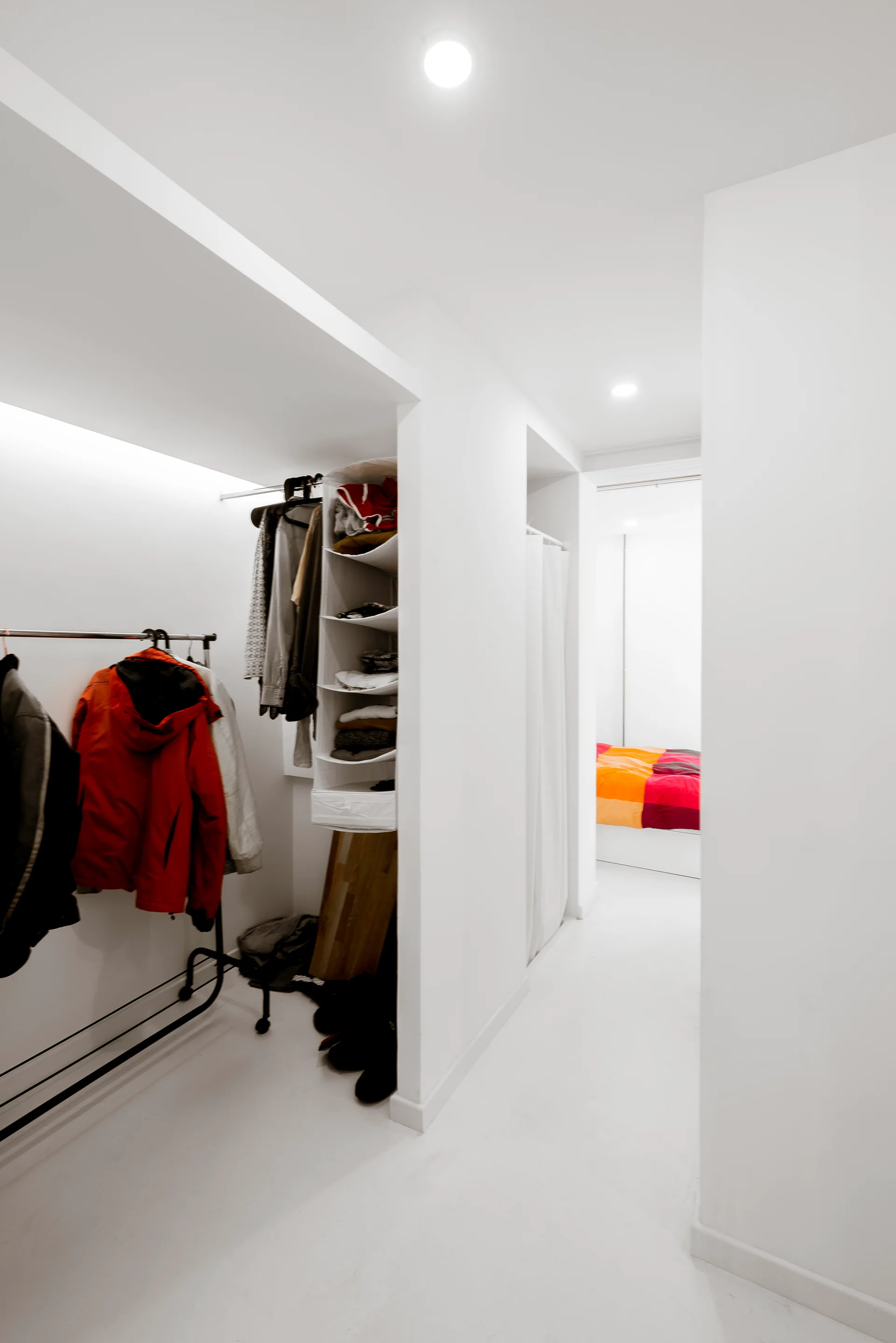 A narrow, minimalist walk-in closet with white walls and floors features an open metal clothing rack and hanging fabric storage shelves.