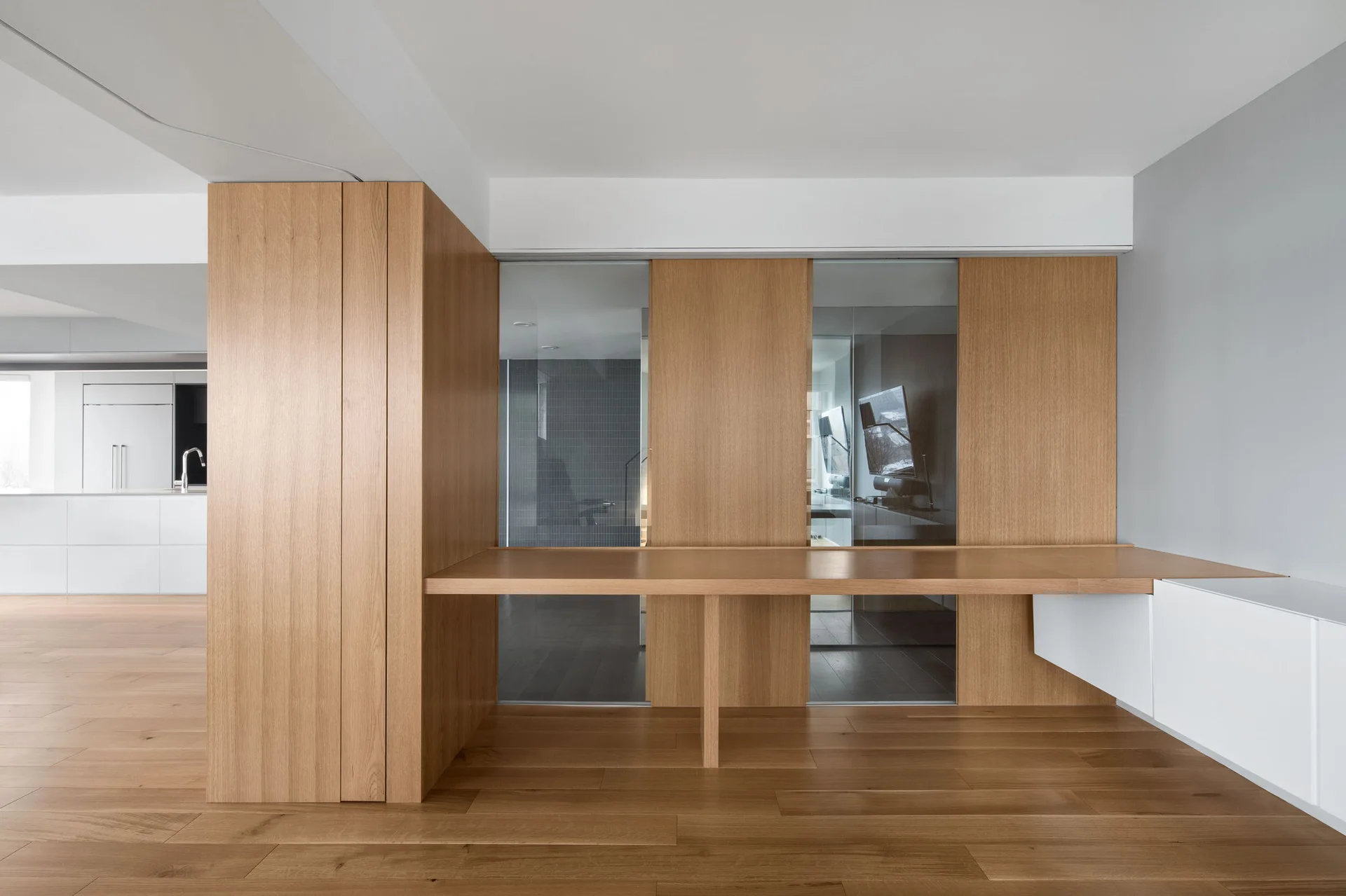 A minimalist study featuring a custom oak built-in desk integrated into matching wood wall panels with glass partitions and white storage cabinetry.