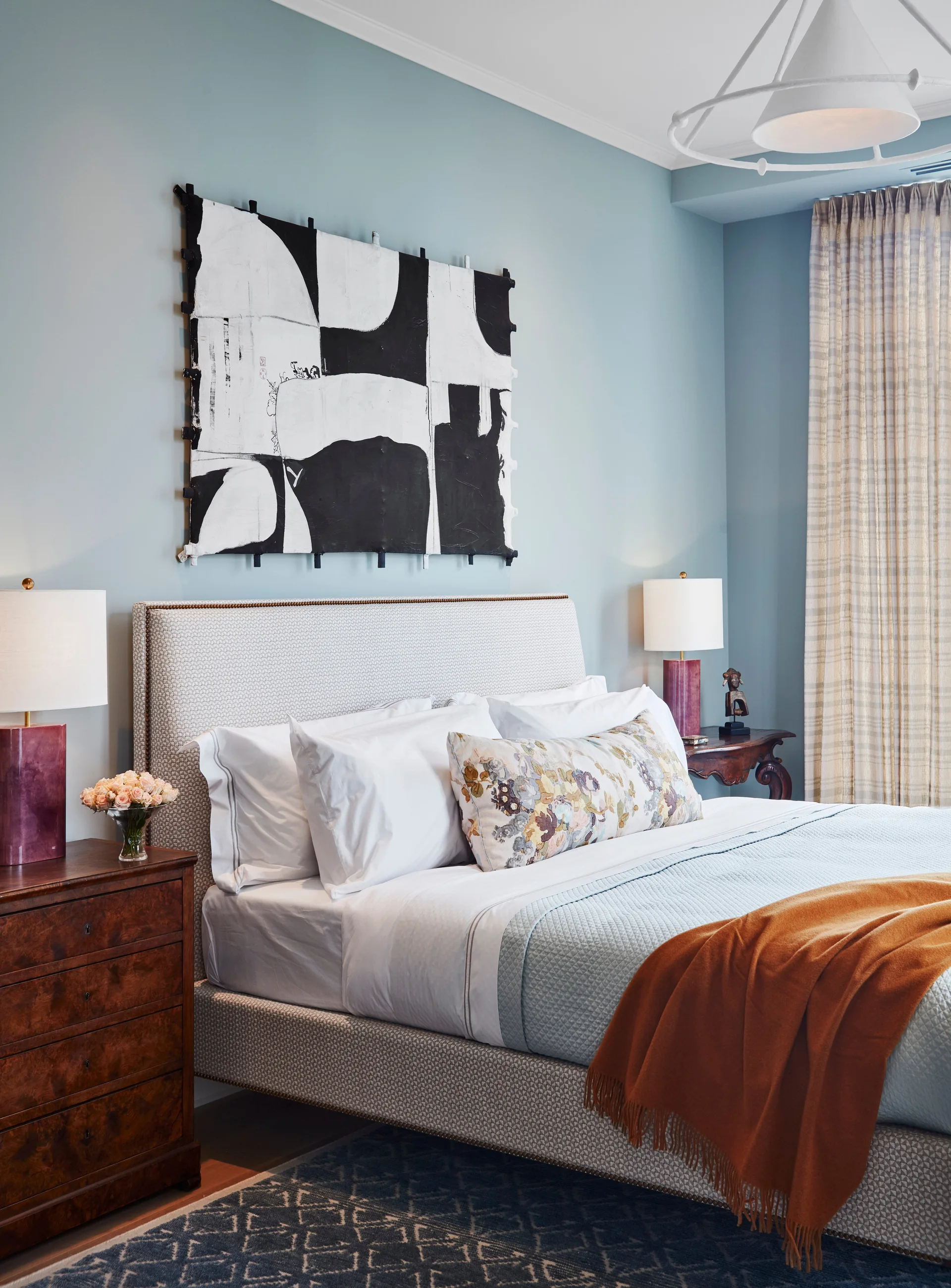 A bedroom features a light blue wall adorned with abstract black and white art above an upholstered bed with a patterned headboard.