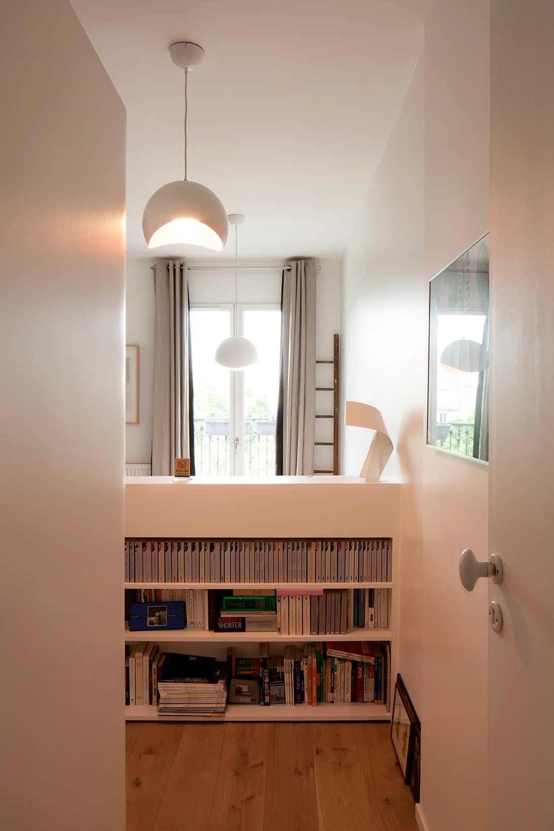 View through a narrow white hallway showing a built-in bookshelf partition, light wood floors, and white dome pendant lights hanging from the ceiling.