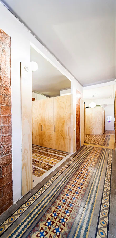A long hallway features intricate geometric patterned floor tiles, light plywood wall panels, a built-in closet, and an exposed brick accent wall.
