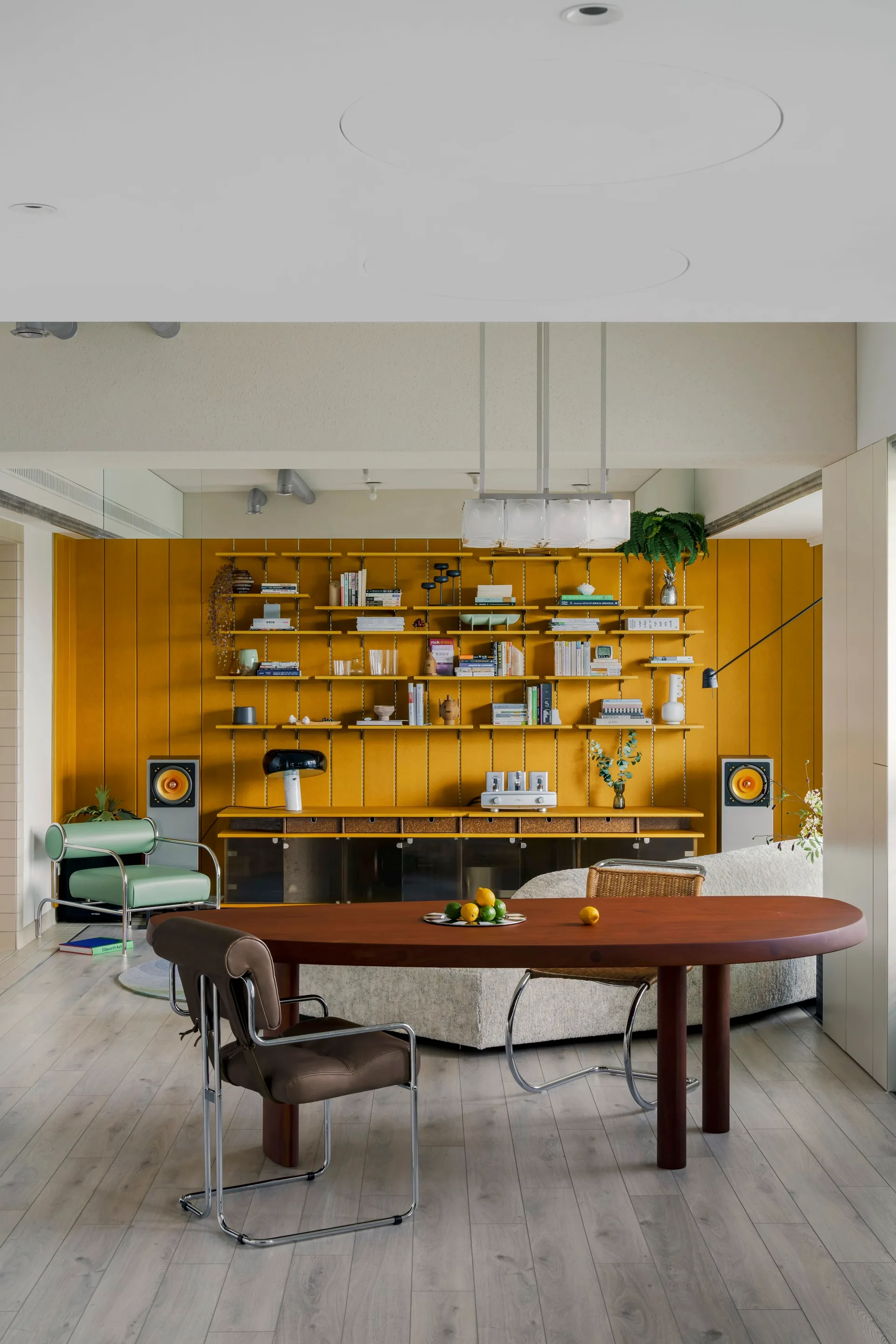 An open living and dining room with a mustard yellow paneled wall, modular shelving, a dark wood oval dining table, and a curved cream sofa.