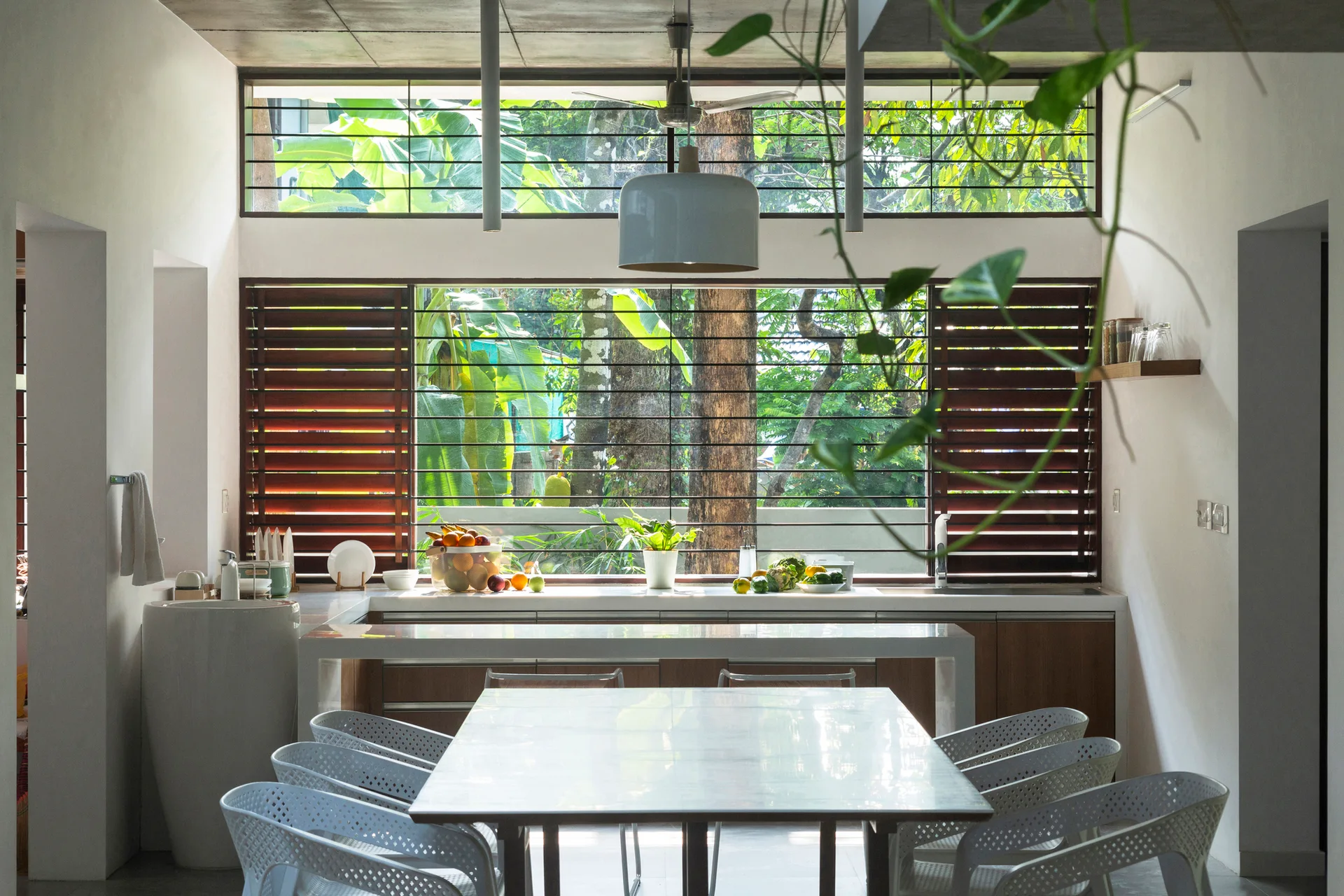 A modern dining space with a white table and chairs sits before a kitchen counter and a large shuttered window framing lush green trees.