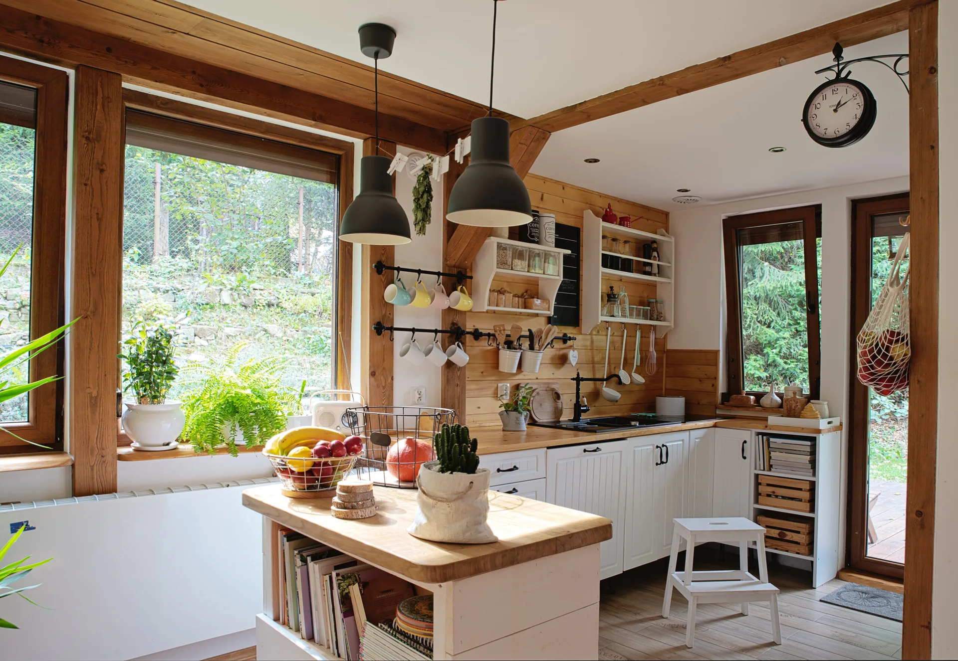 A rustic kitchen features white base cabinets, wooden countertops, exposed timber beams, a central kitchen island, and large windows.