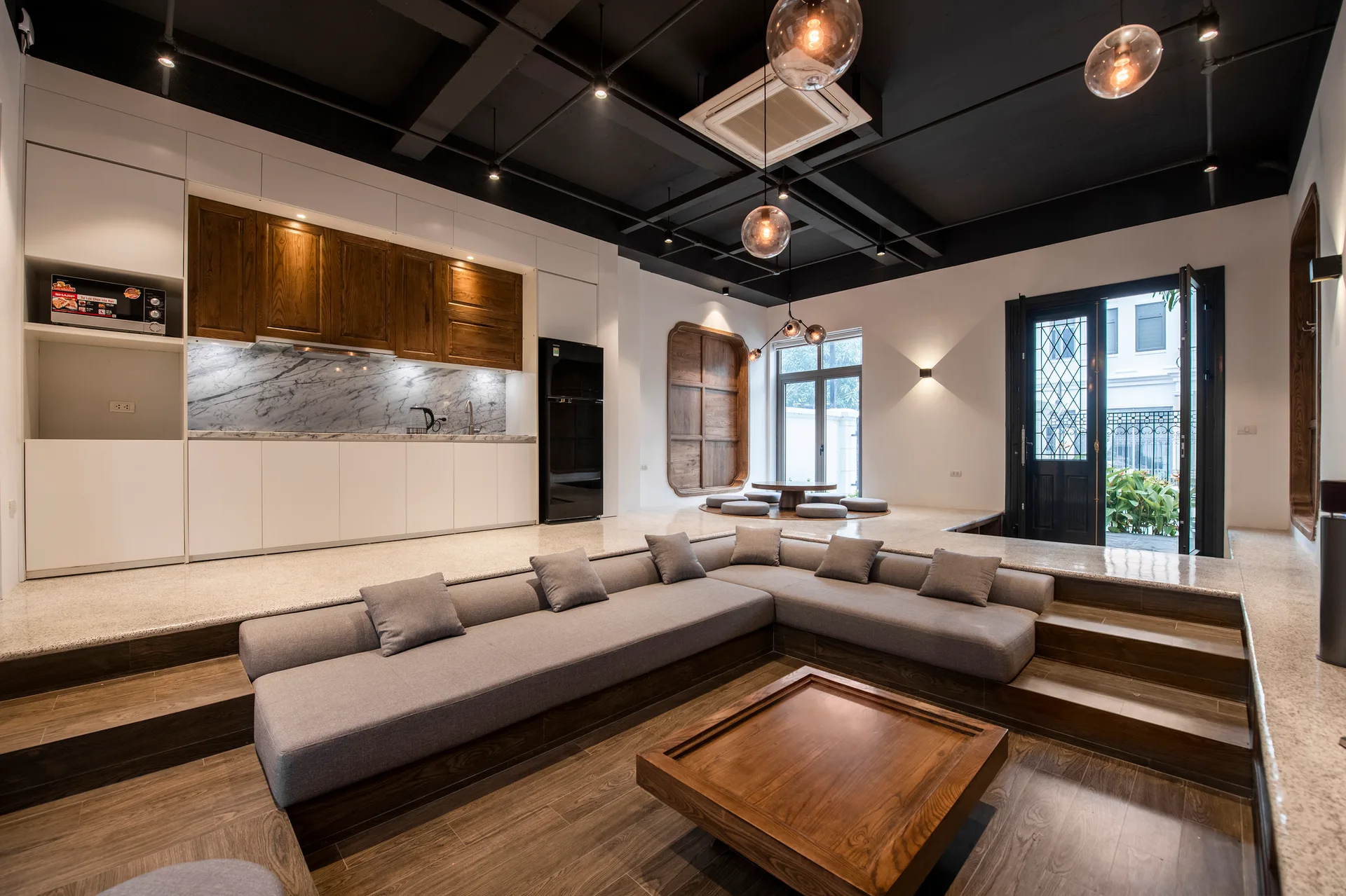 An open-concept room features a sunken grey sectional sofa, a low wooden coffee table, a black exposed ceiling, and a modern kitchen.