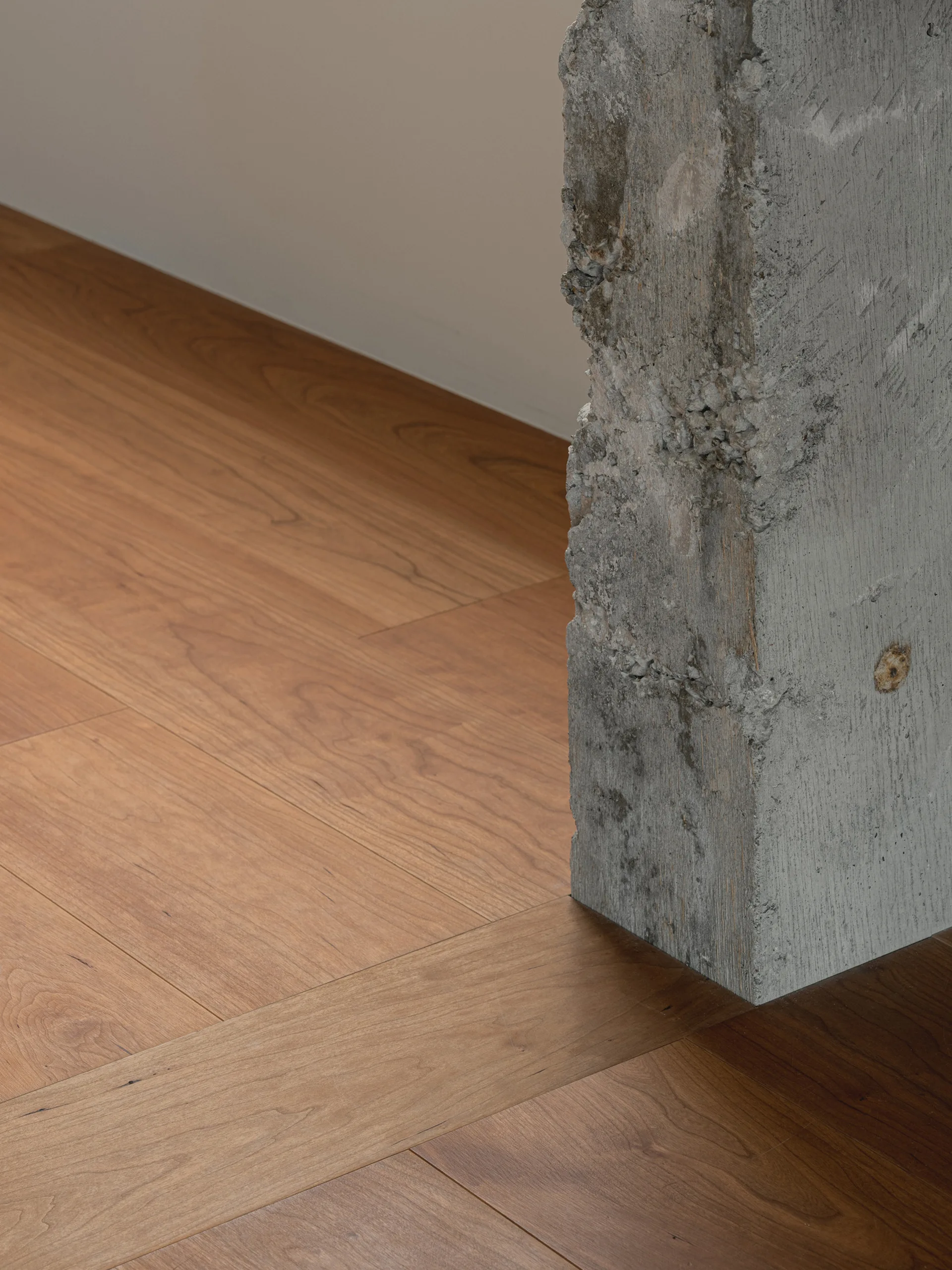 A close-up view of a rough, unfinished gray concrete column where it meets smooth, warm brown wood plank flooring next to a plain wall.