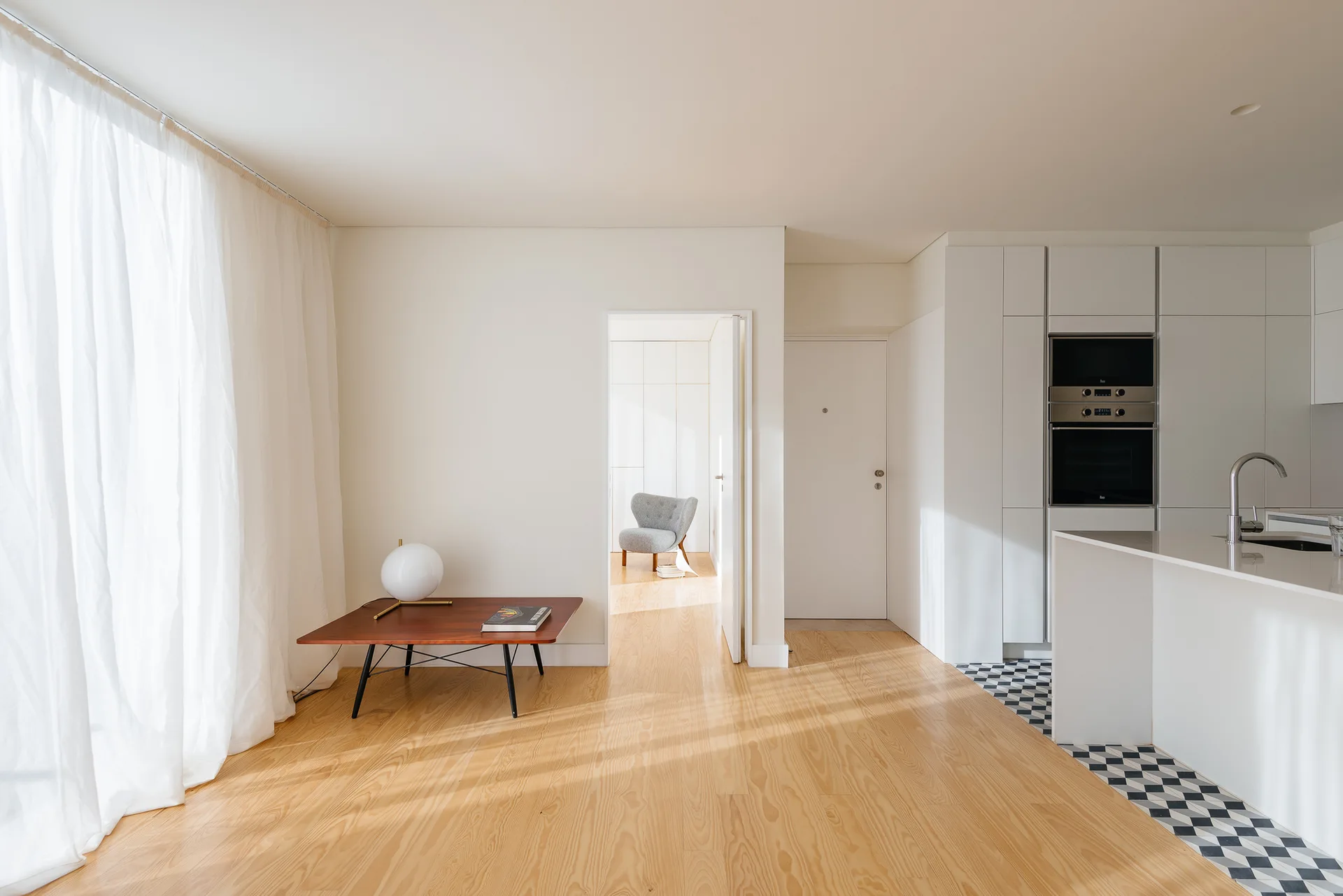 A bright minimalist interior features light wood flooring transitioning to a white kitchen with a checkerboard tile floor and sheer curtains.
