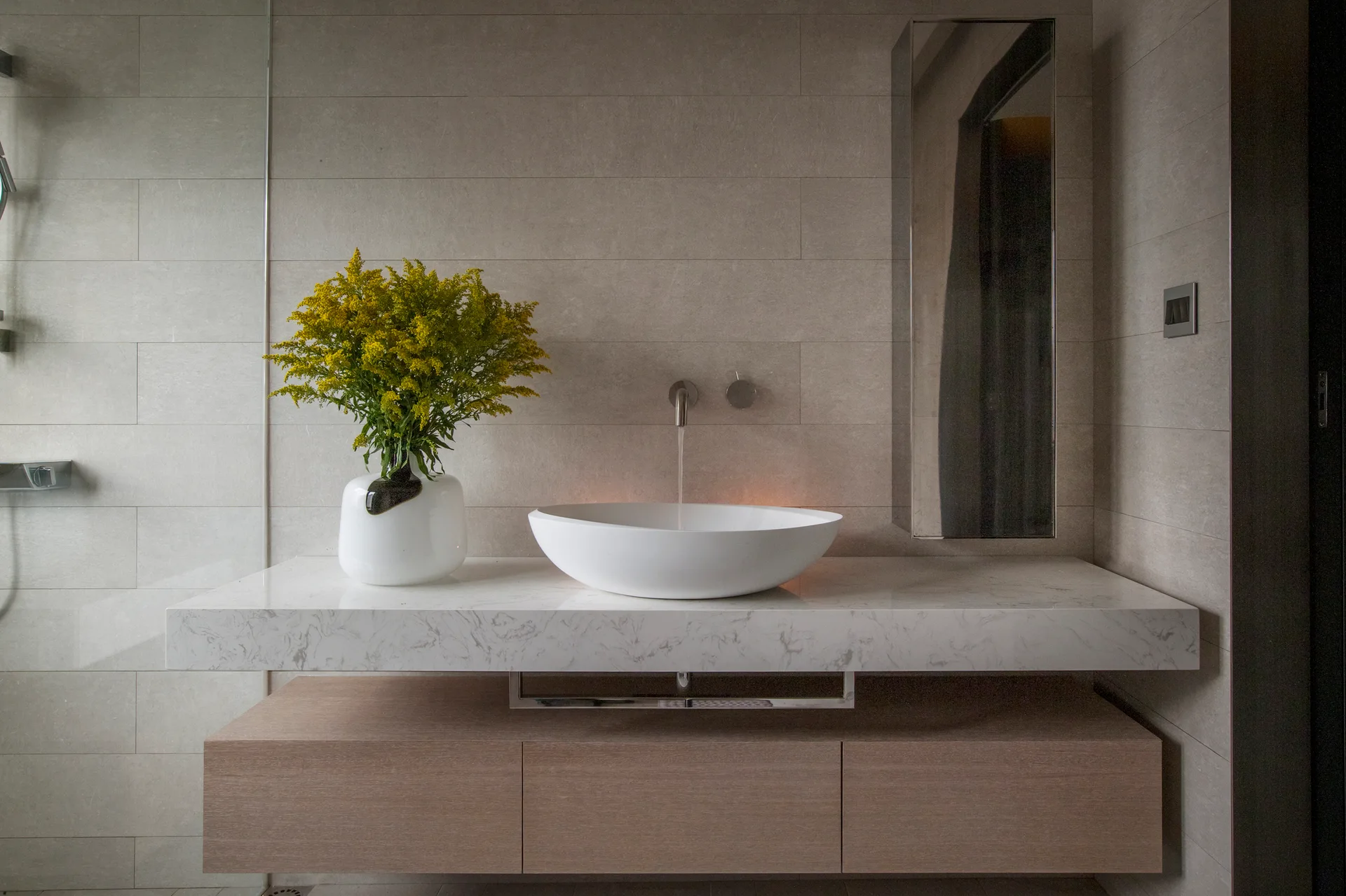 A modern floating vanity features a thick marble-veined countertop, a white oval vessel sink, a light wood lower cabinet, and beige wall tiles.