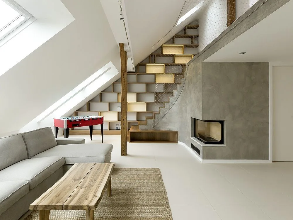 A modern loft living room features a grey sectional sofa, sloped ceiling skylights, a concrete fireplace, and a wooden staircase with built-in shelving.