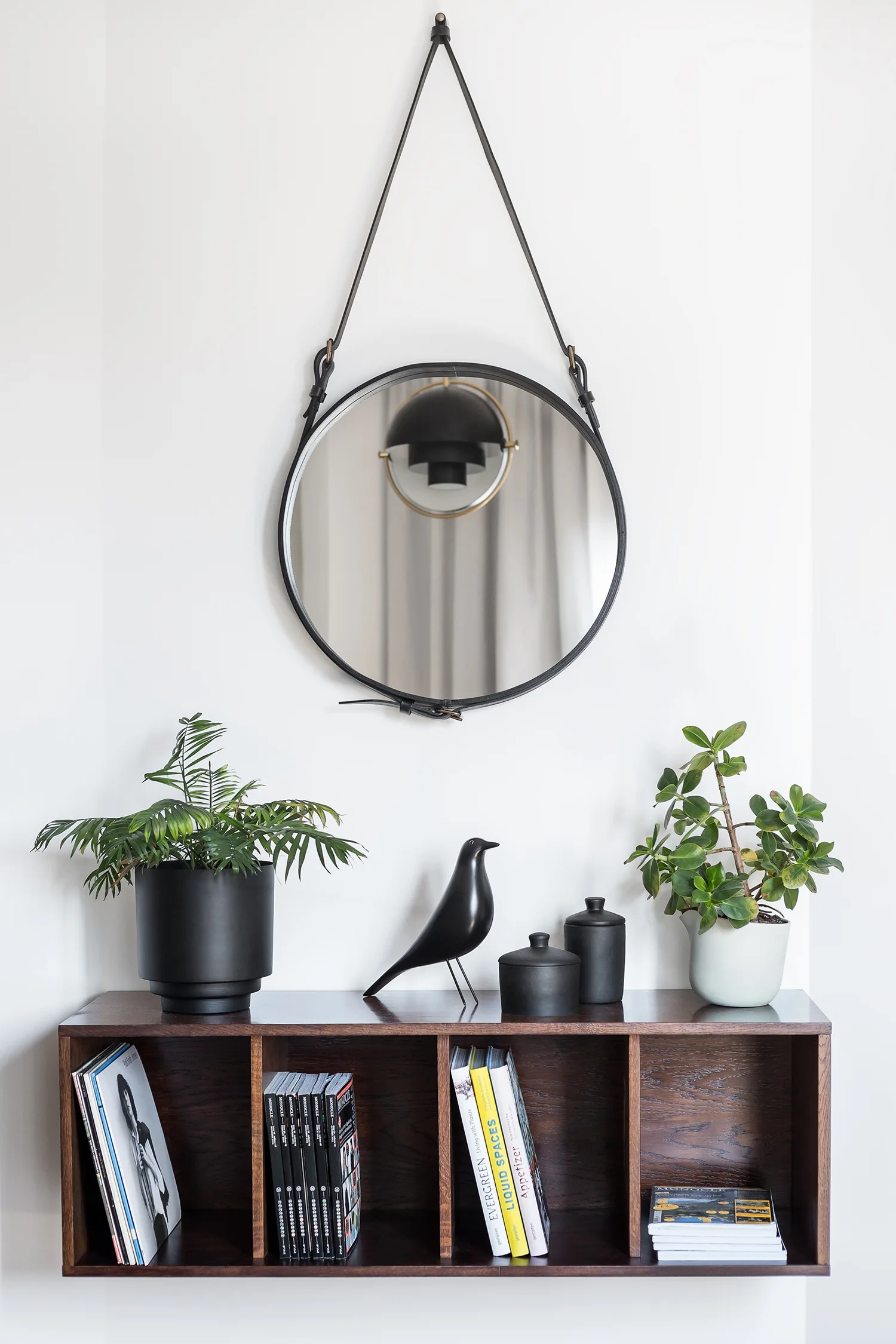 A circular mirror suspended by a black leather strap hangs on a white wall above a dark brown wood cubby shelf styled with books and plants.
