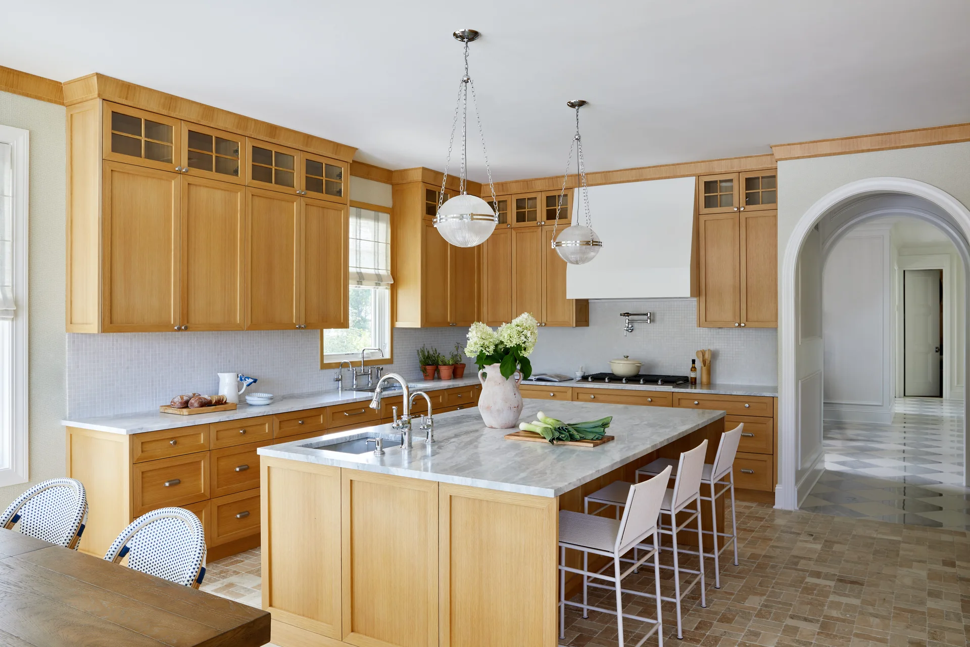 A bright kitchen features warm wood cabinetry, a grey-veined marble island with white stools, spherical pendant lights, and an arched doorway.