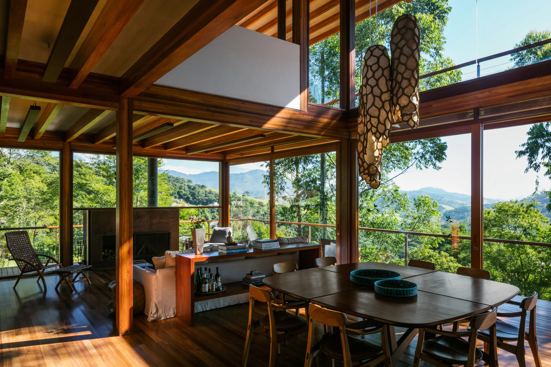 An open-concept living and dining room featuring structural wood framing, a large wooden table with chairs, and glass walls overlooking green hills.
