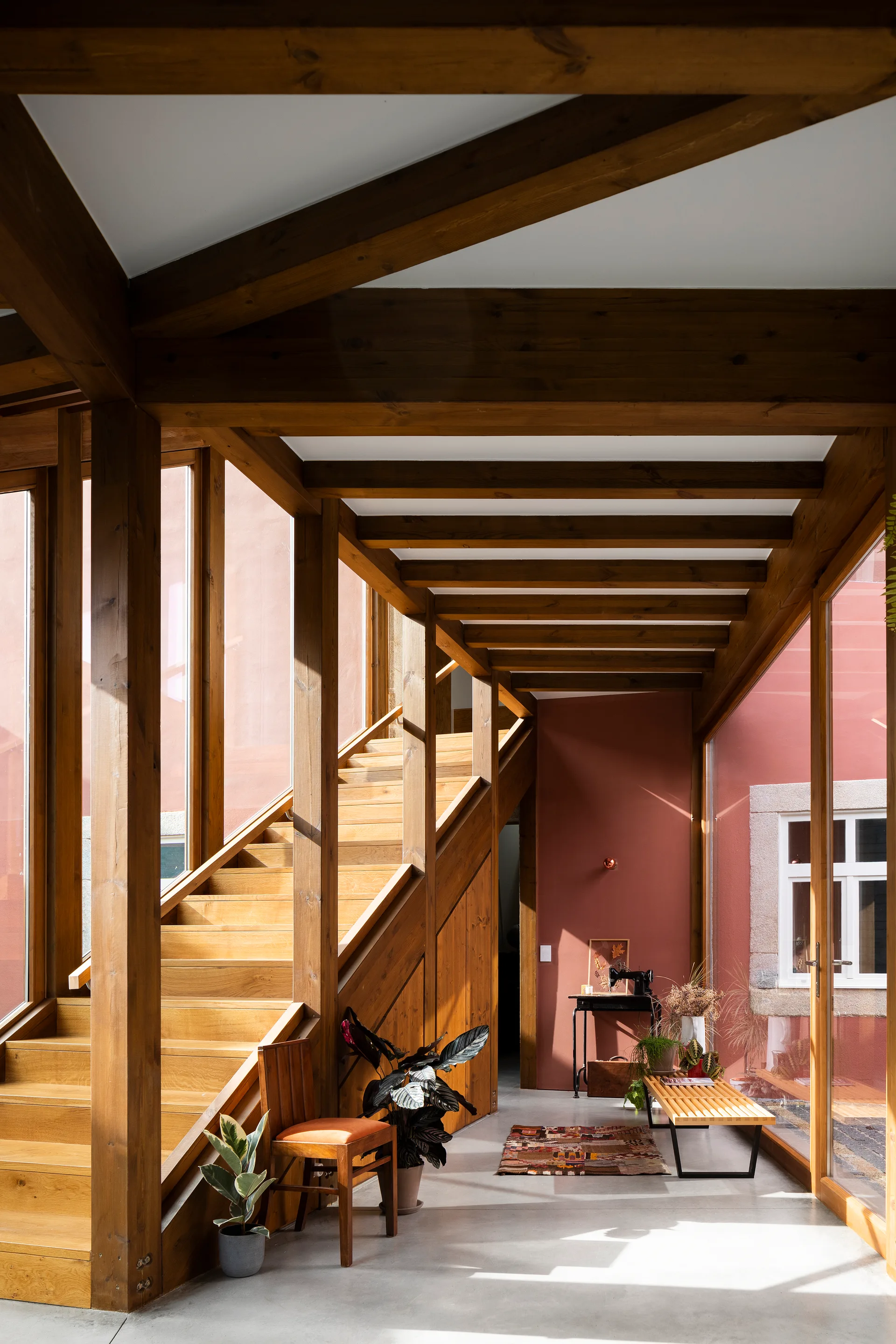 A bright entryway features a wooden staircase, exposed timber ceiling beams, concrete flooring, and a deep red accent wall next to glass windows.