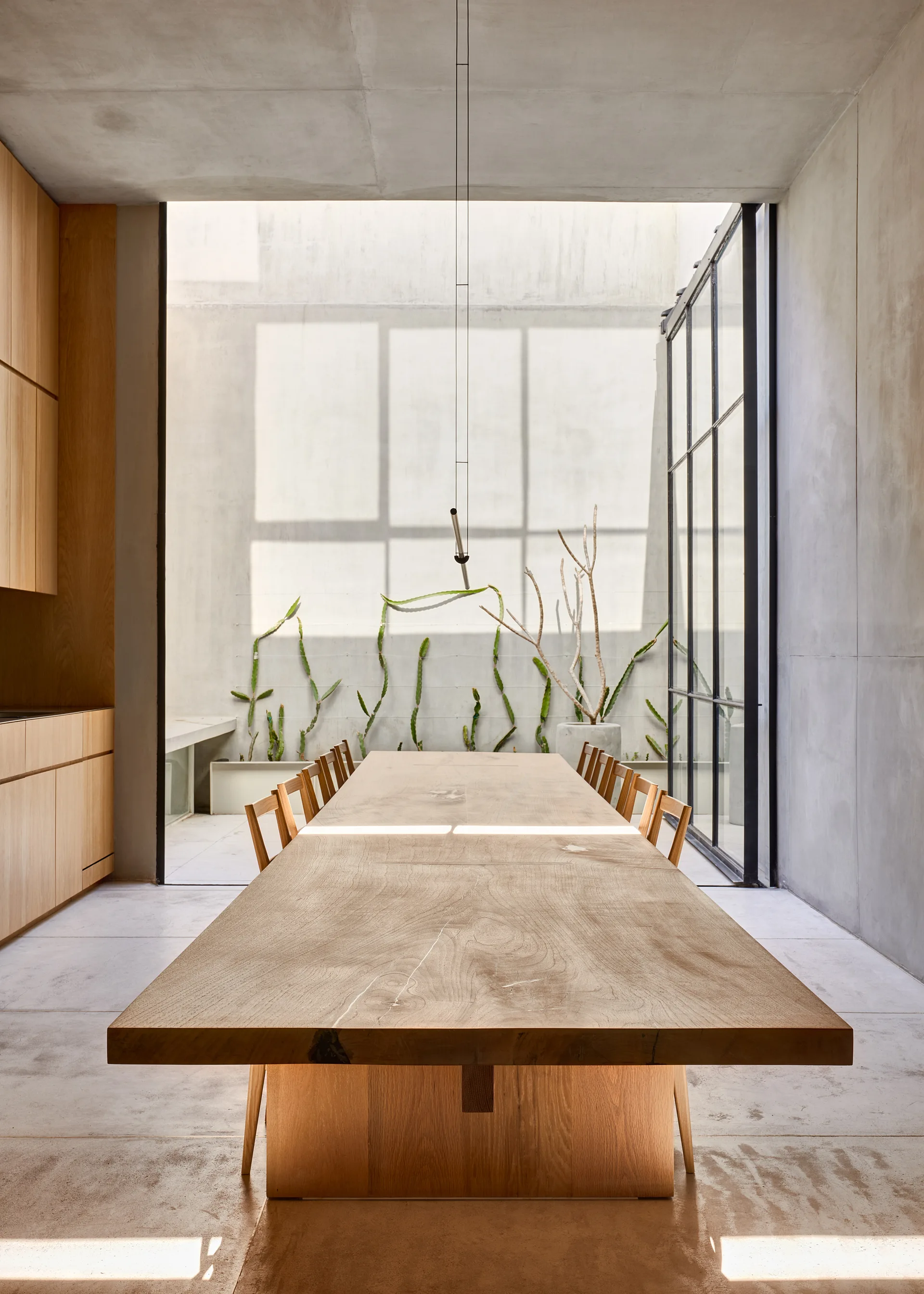 A long wooden dining table and chairs sit in a concrete room next to a glass door opening to a sunlit courtyard with plants.