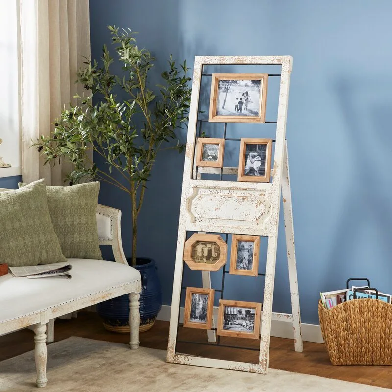 A tall distressed white wood photo collage frame stands on a wooden floor next to an upholstered bench and olive tree planter against a blue wall.