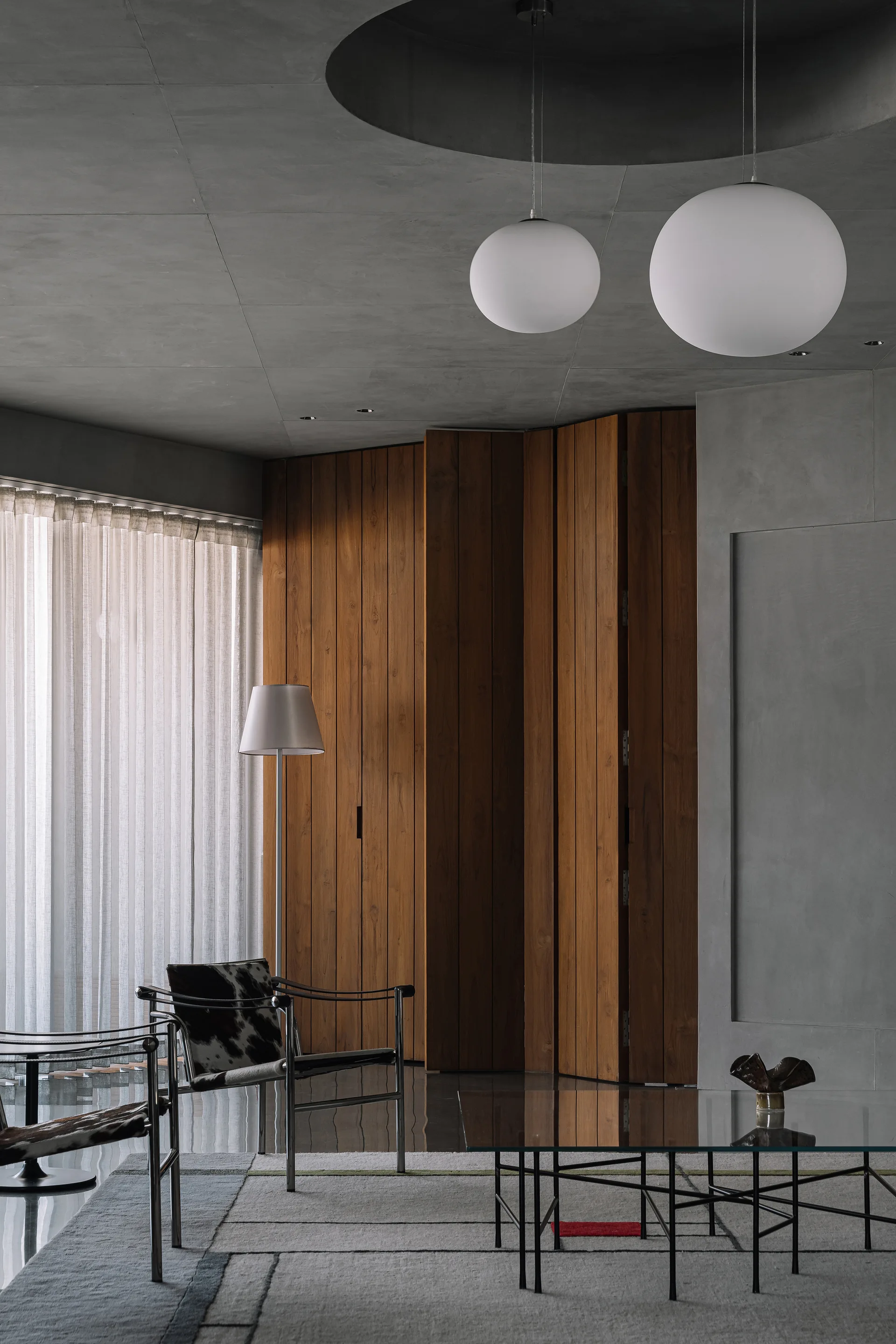 A modern living room features a grey concrete ceiling, a folding wood-paneled wall, globe pendant lights, and two cowhide chairs on a geometric rug.
