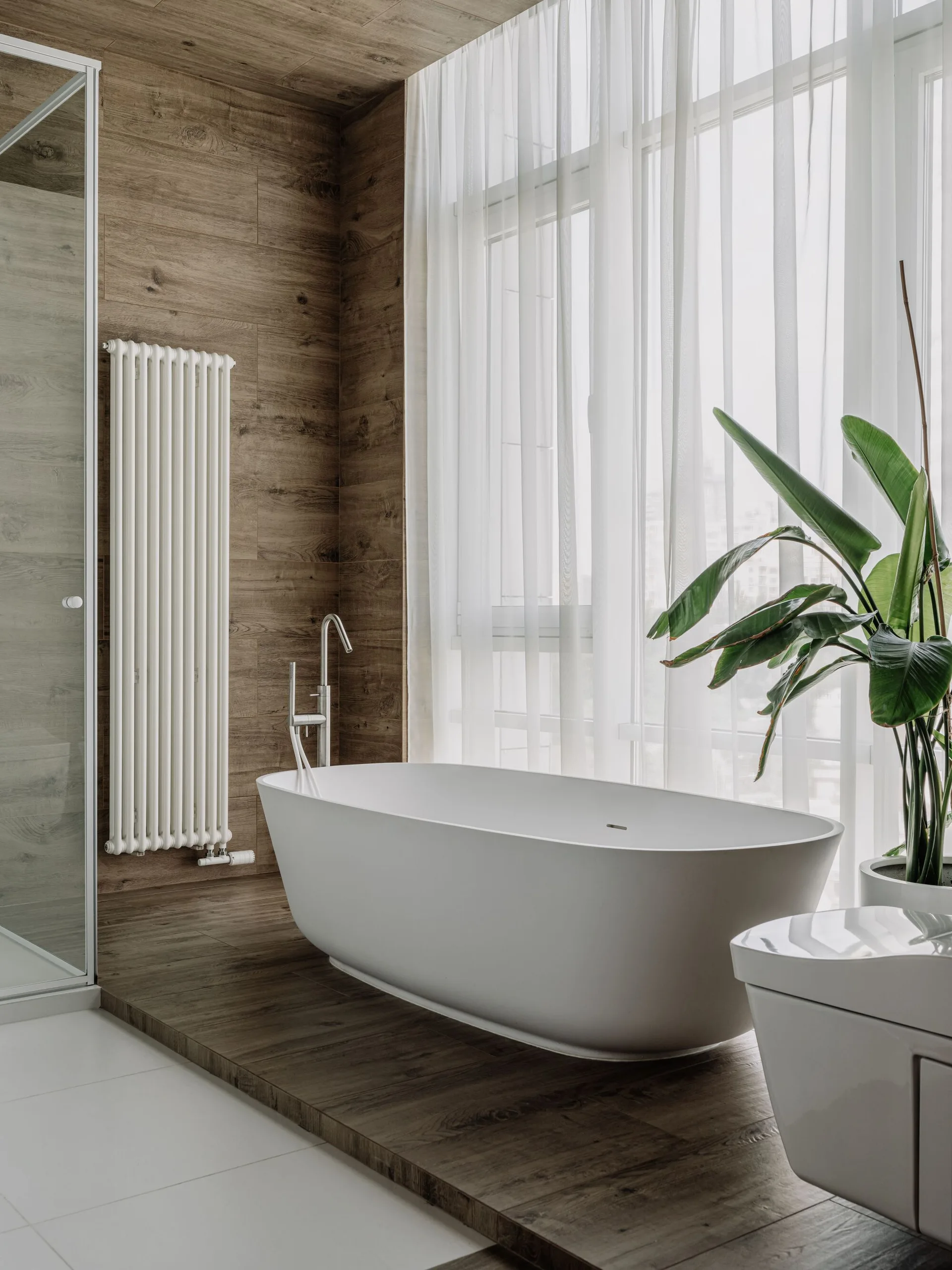 A white freestanding bathtub sits on a raised wood-plank platform next to a tall window with sheer white curtains and a vertical radiator.