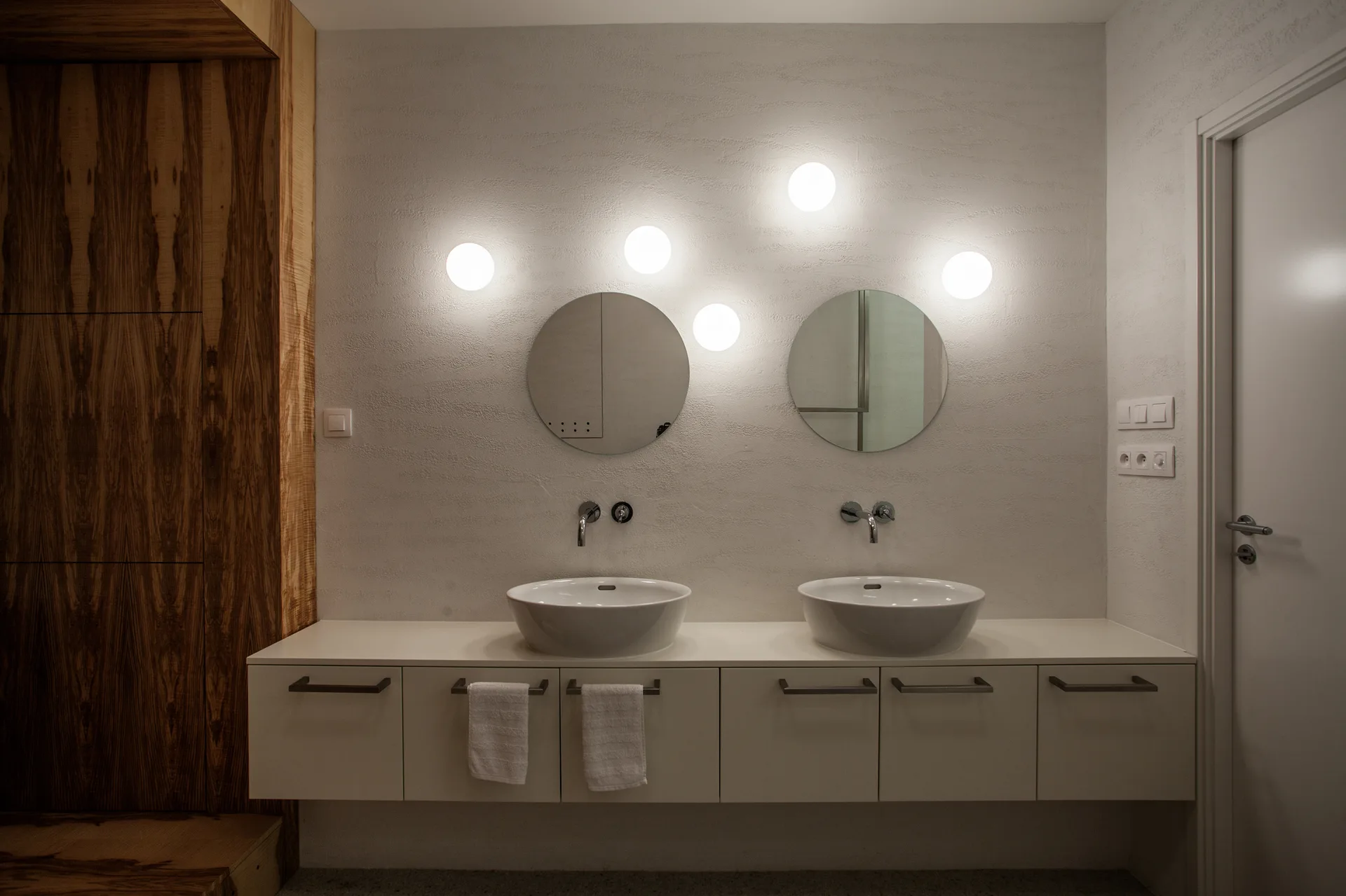 A minimalist double bathroom vanity featuring white vessel sinks, round mirrors, and five glowing orb sconces against a textured plaster wall.