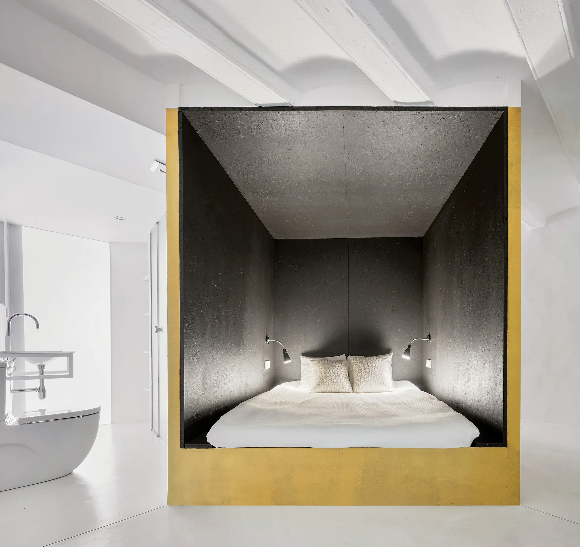 A bed sits inside a black box alcove with a gold border set against white walls, a white floor, and exposed ceiling beams.
