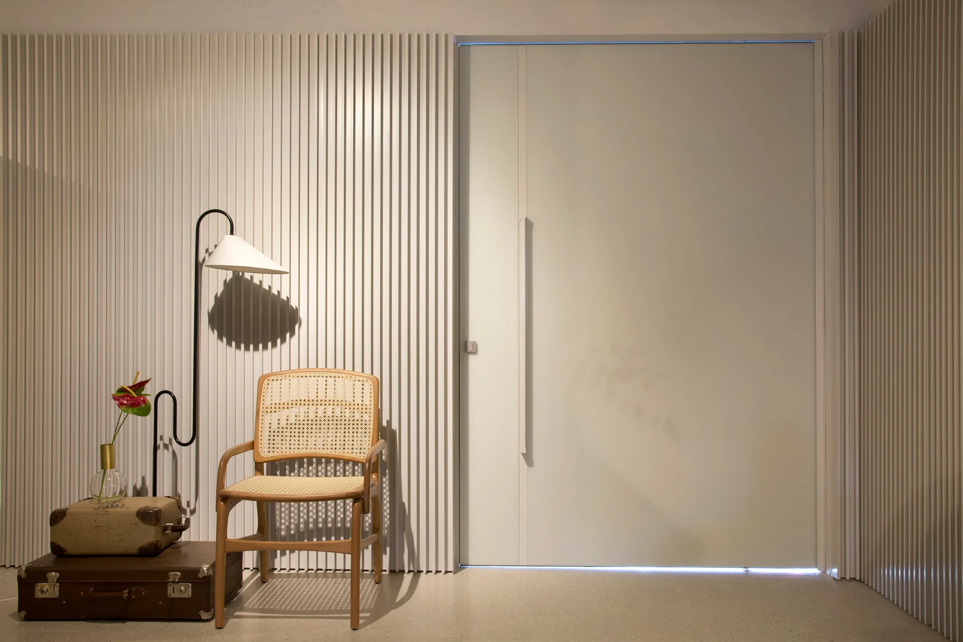 A minimalist entryway featuring white vertical slatted wall paneling, a flush door, a wooden cane chair, and stacked vintage suitcases.