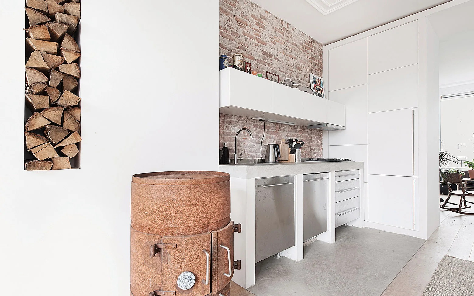 A modern kitchen features white minimalist cabinets, an exposed brick wall, a vertical firewood alcove, and a rust-colored cylindrical stove.
