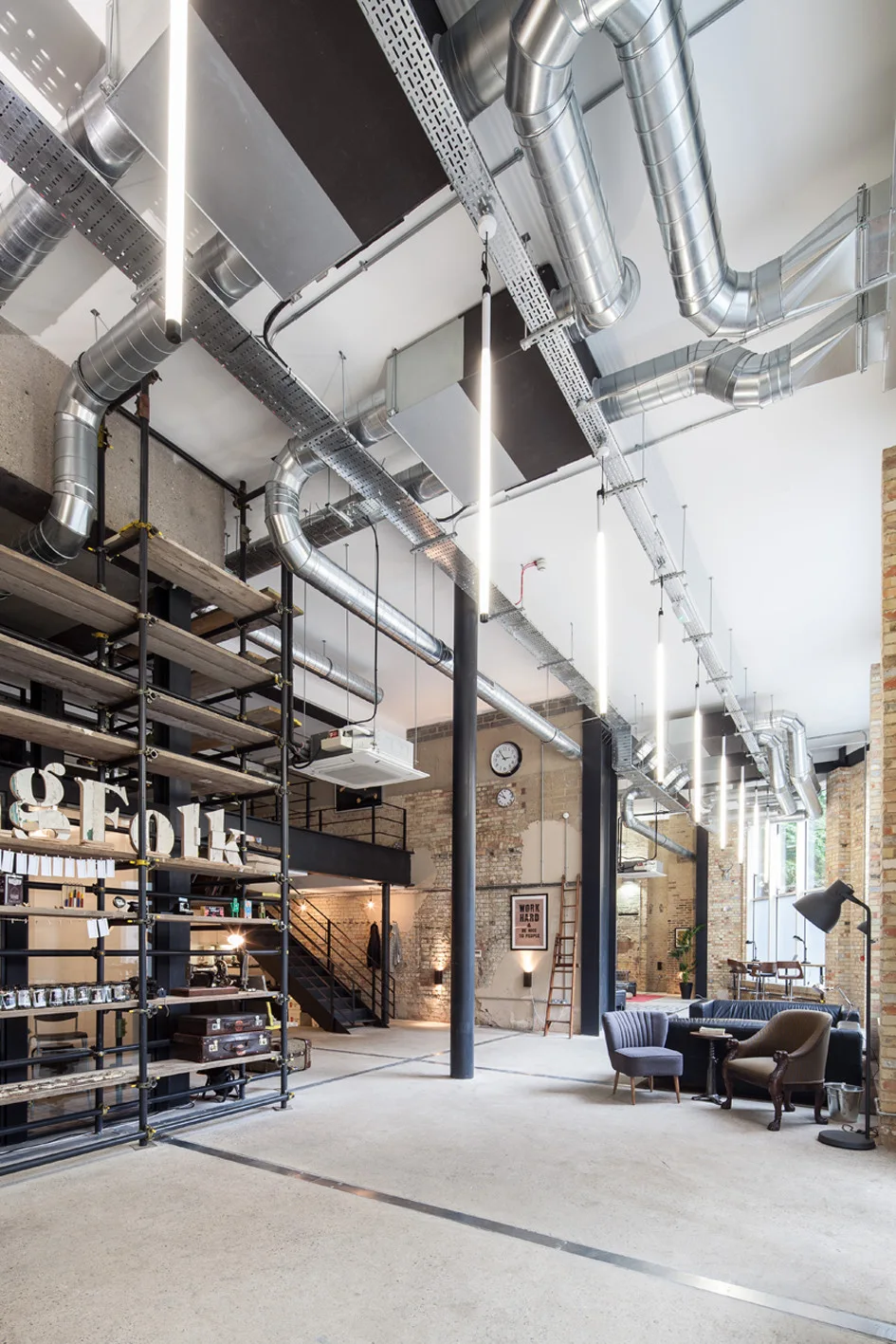 A spacious industrial loft with exposed ceiling ducts, brick walls, polished concrete floors, suspended tube lights, and a scaffold shelving unit.