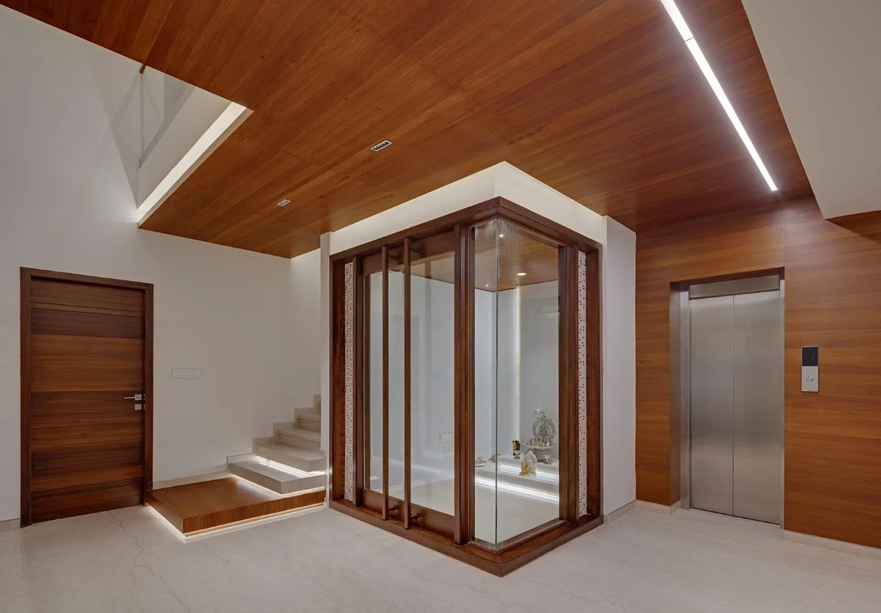 A modern residential entryway featuring warm wood ceiling paneling, a glass-enclosed central shrine, stone steps, and an integrated stainless steel elevator.