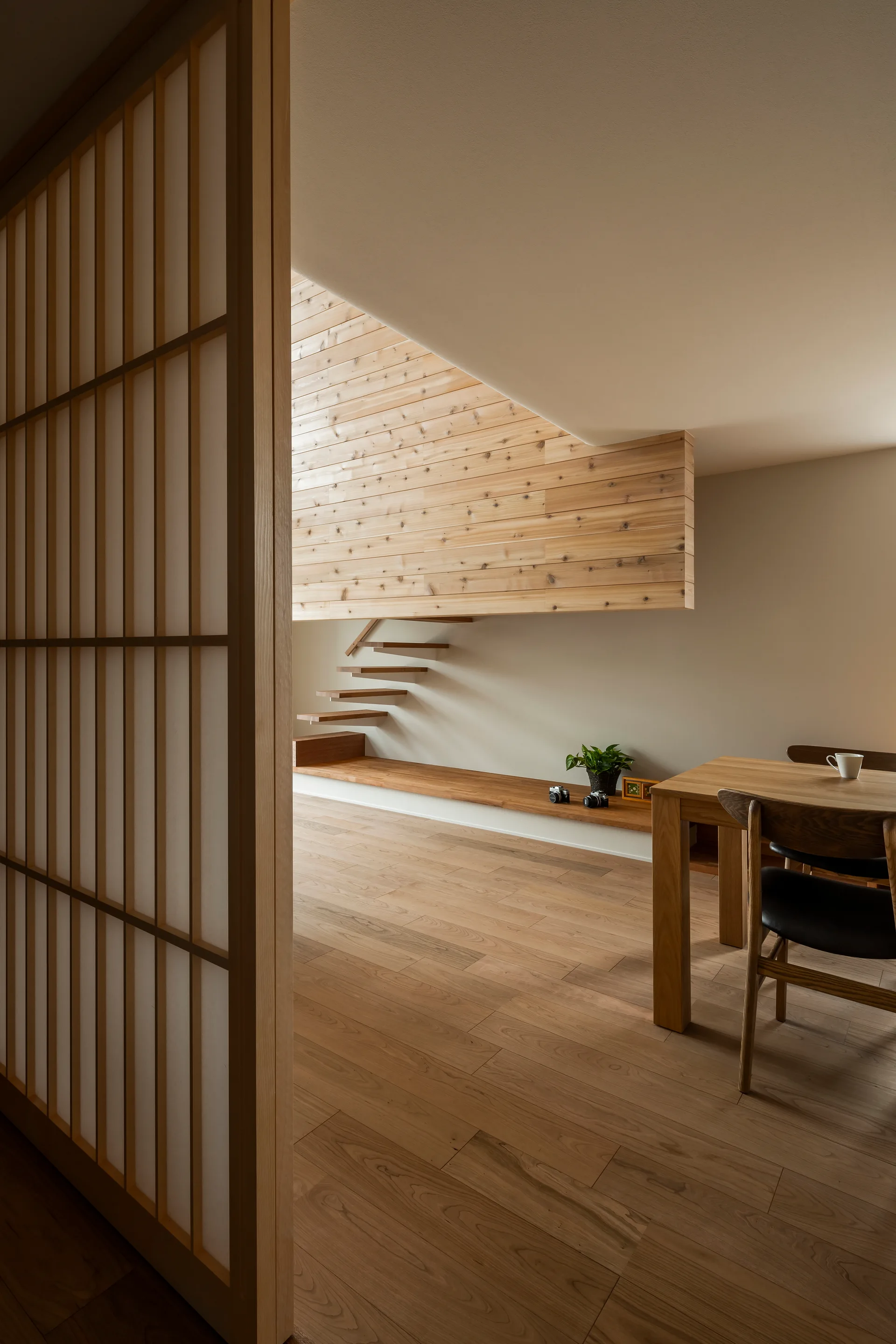 A minimalist dining room features a wooden table and chairs, a sliding shoji screen, and a cantilevered floating staircase against a wood-paneled wall.