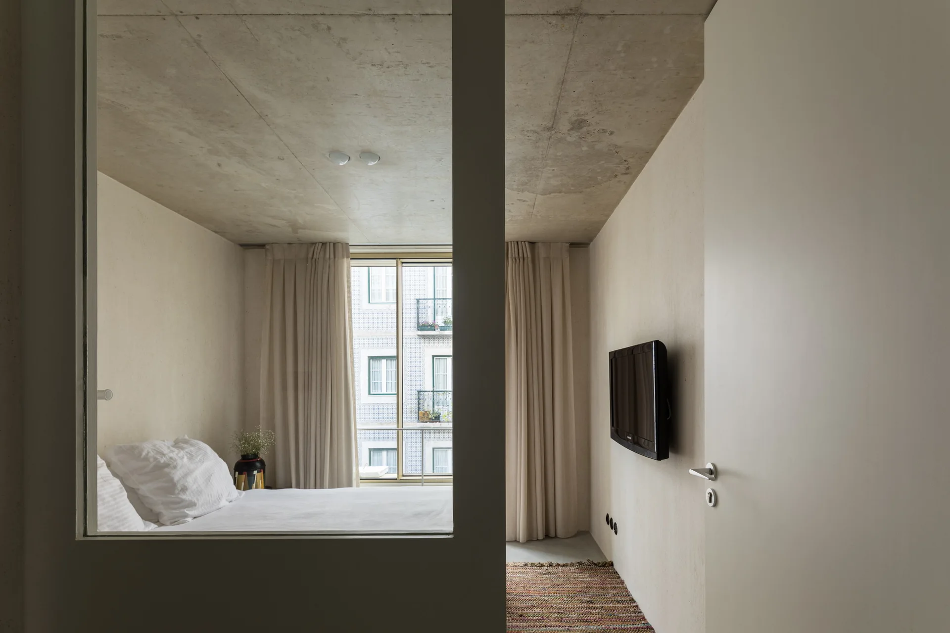 A minimalist bedroom with a raw concrete ceiling, beige plaster walls, and a bed with white bedding, viewed through an interior wall cutout.