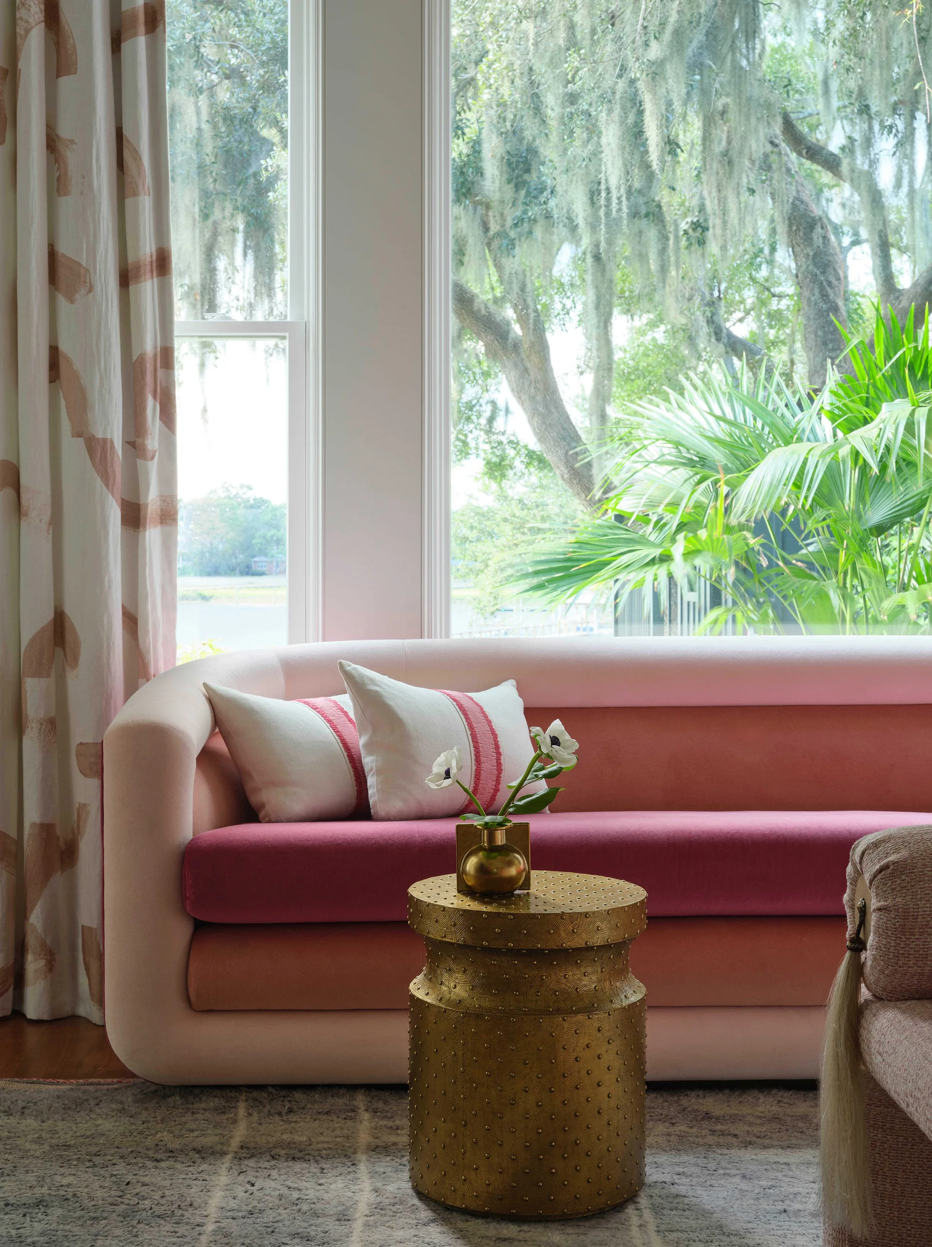 A multi-toned pink sofa with striped throw pillows sits in front of a large window next to a patterned curtain and a textured gold side table.