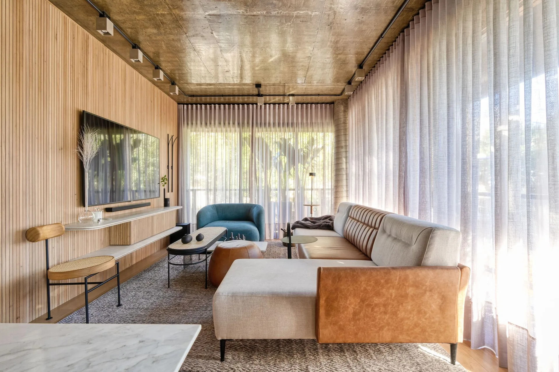Modern living room with a vertical wood slat wall, a modular leather and fabric sofa, sheer curtains, and an exposed concrete ceiling.