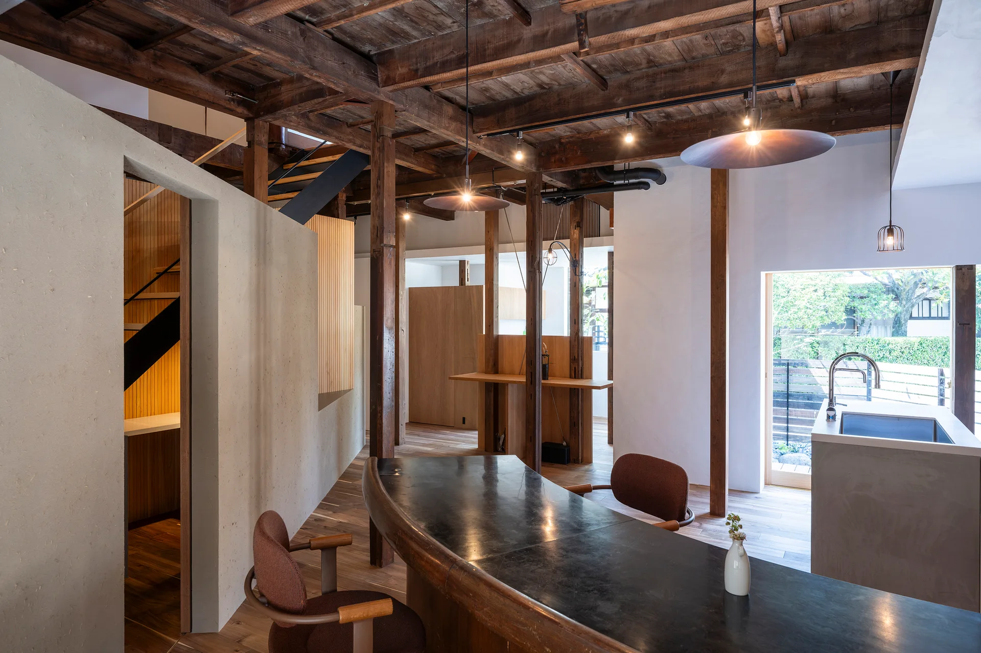 A modern dining space features a dark curved table, two fabric chairs, and an open kitchen with exposed wooden ceiling beams.