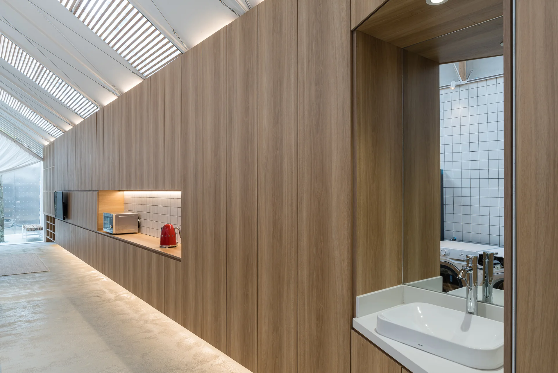 A long wood-paneled storage wall features recessed niches with a kitchen counter and a bathroom vanity under a slanted louvered skylight.