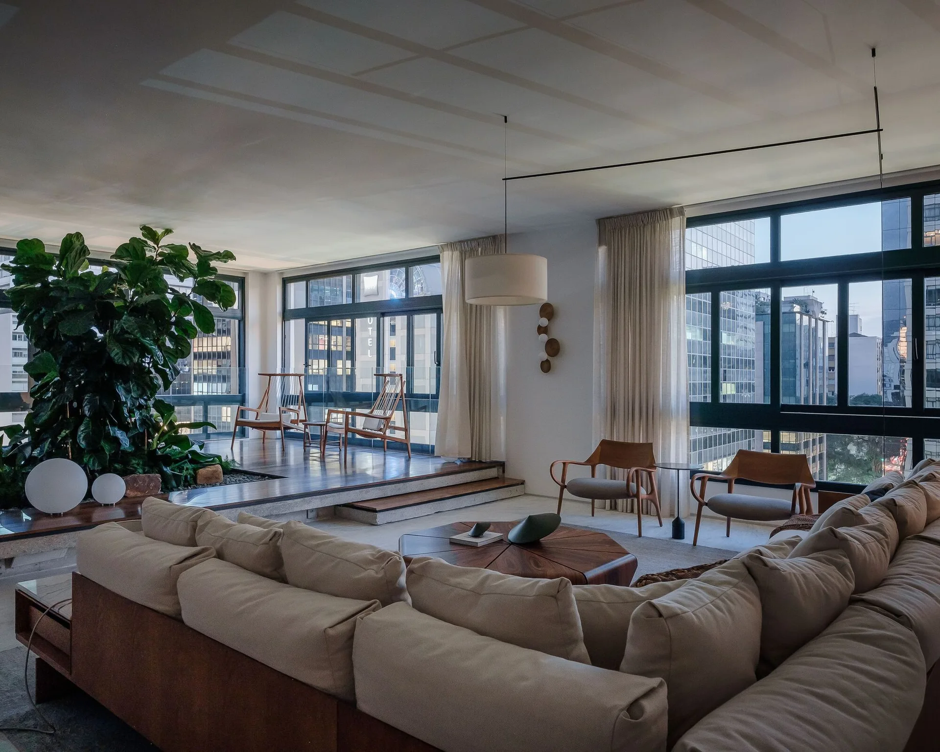 A modern living room features a large beige sectional sofa, a round wood coffee table, and a raised wooden platform area by large windows.