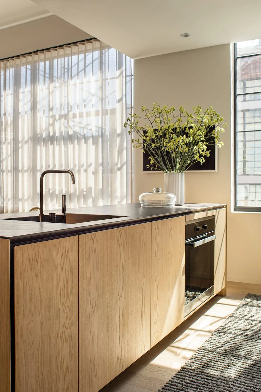 A light wood kitchen island features a dark countertop with a sink, an integrated oven, and a fluted vase with green branches next to sheer curtains.
