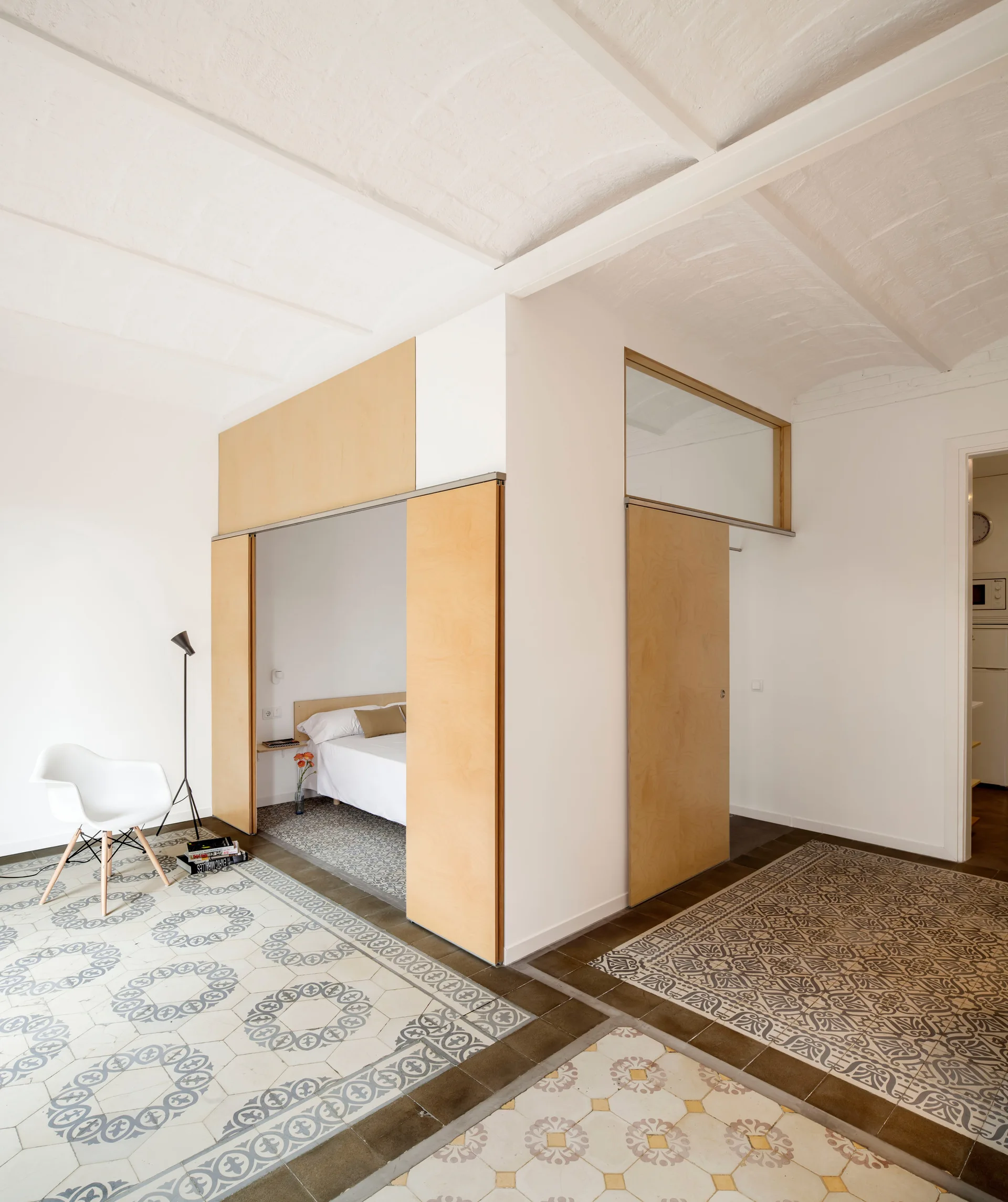 A minimalist bedroom alcove features light wood sliding partition doors, intricate patterned floor tiles, and a white vaulted ceiling.