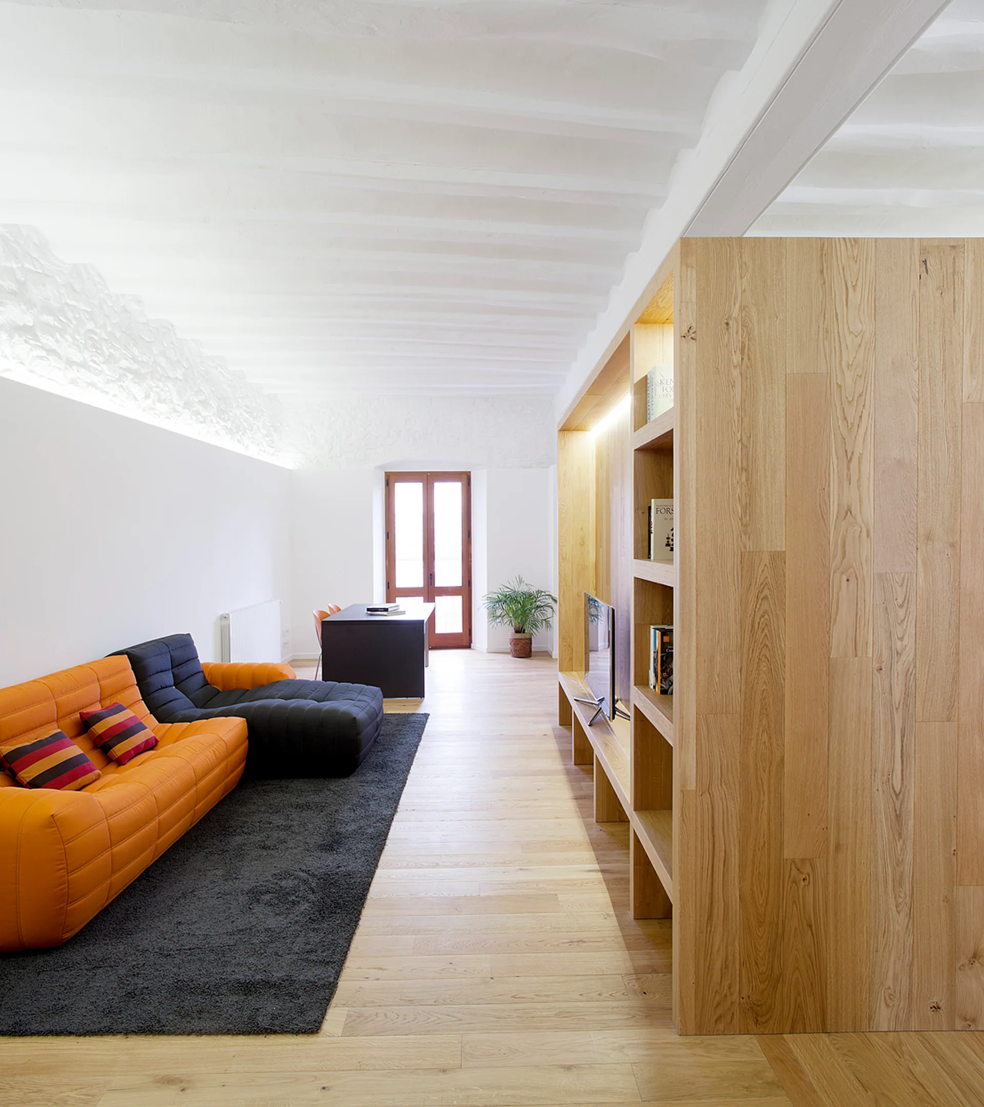 A modern living room features a bright orange and black modular sofa, a dark grey rug, and a large light-wood built-in shelving unit.