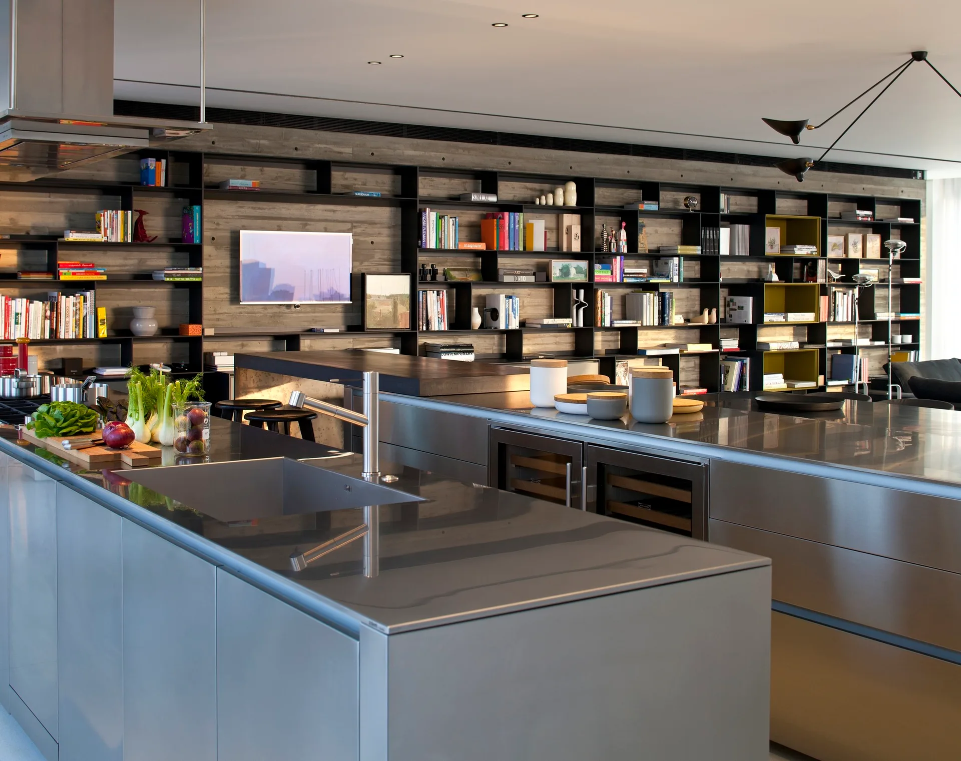 A modern industrial kitchen features two stainless steel islands in front of an expansive black metal shelving unit mounted on a textured concrete wall.
