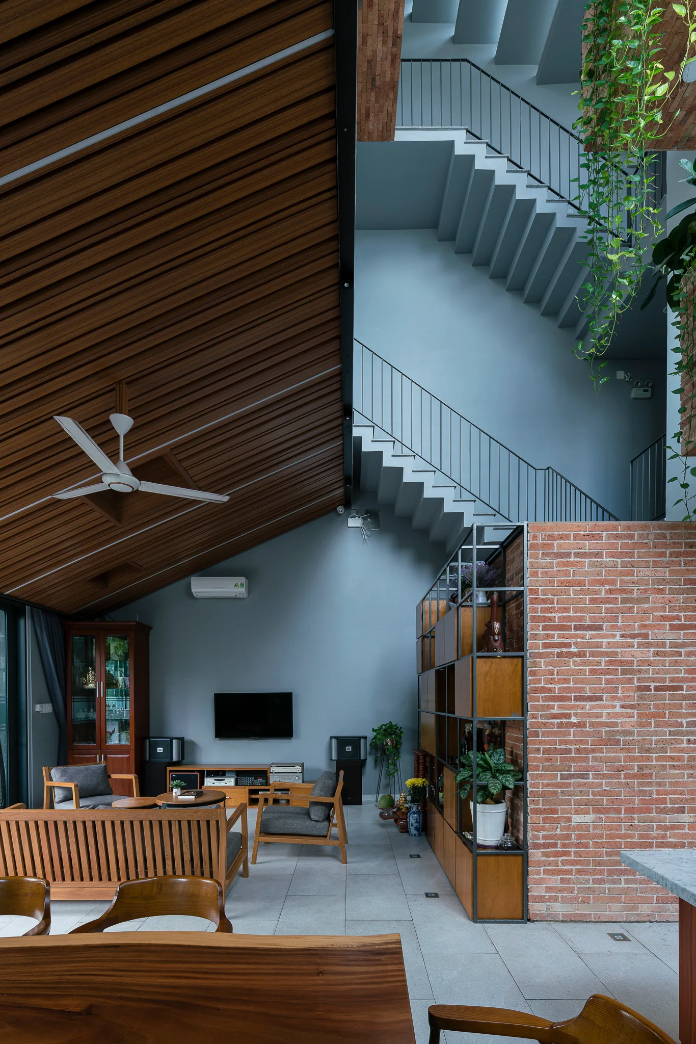 A modern living room features an angled slatted wood ceiling, an open concrete staircase, gray walls, and an exposed brick partition wall.