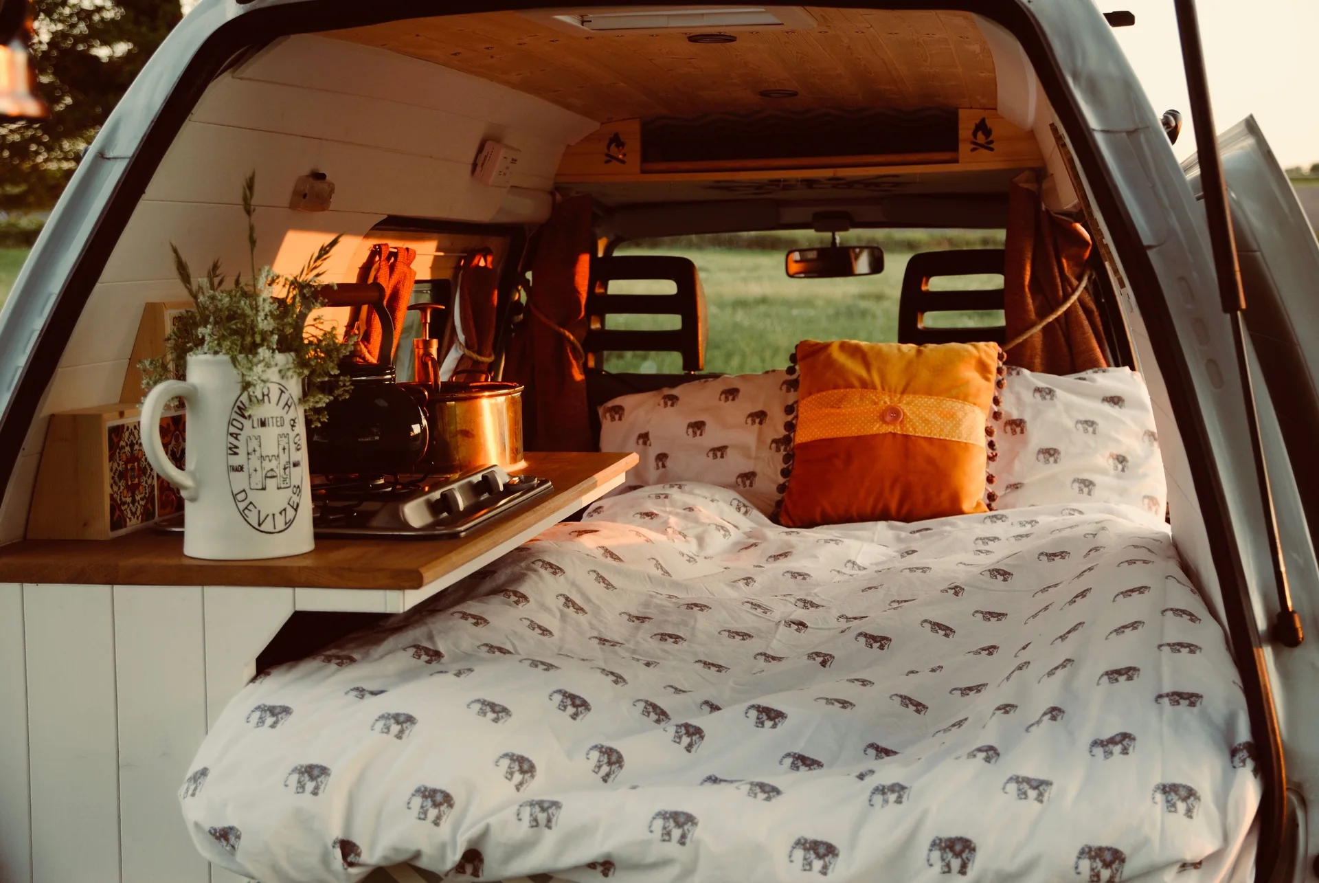 An interior view of a converted camper van featuring a bed with elephant-patterned bedding, an orange pillow, and a small wooden kitchenette.
