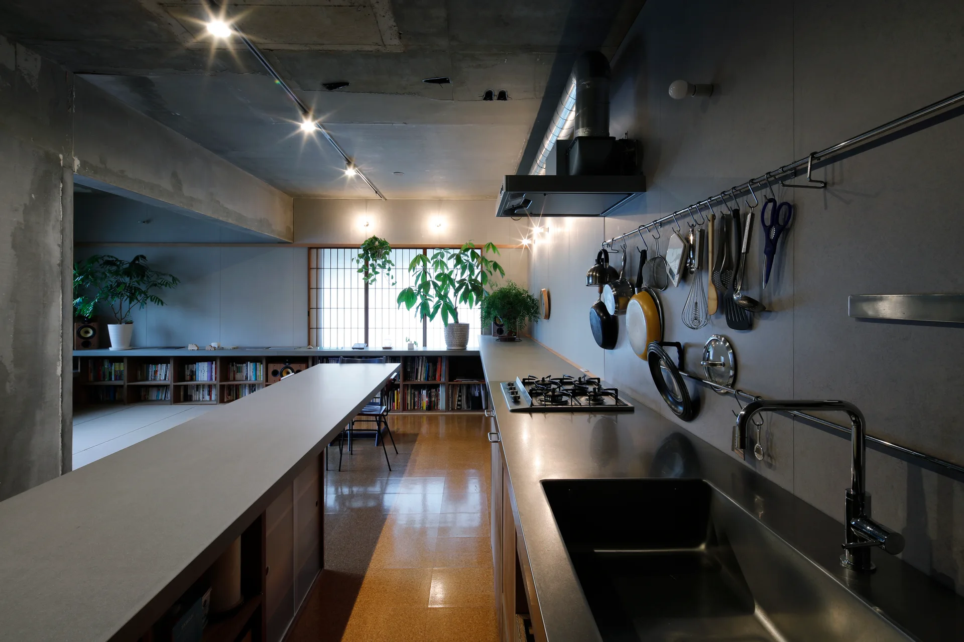 An industrial kitchen featuring a long stainless steel countertop, exposed concrete walls and ceiling, a shoji-style window, and hanging utensils.