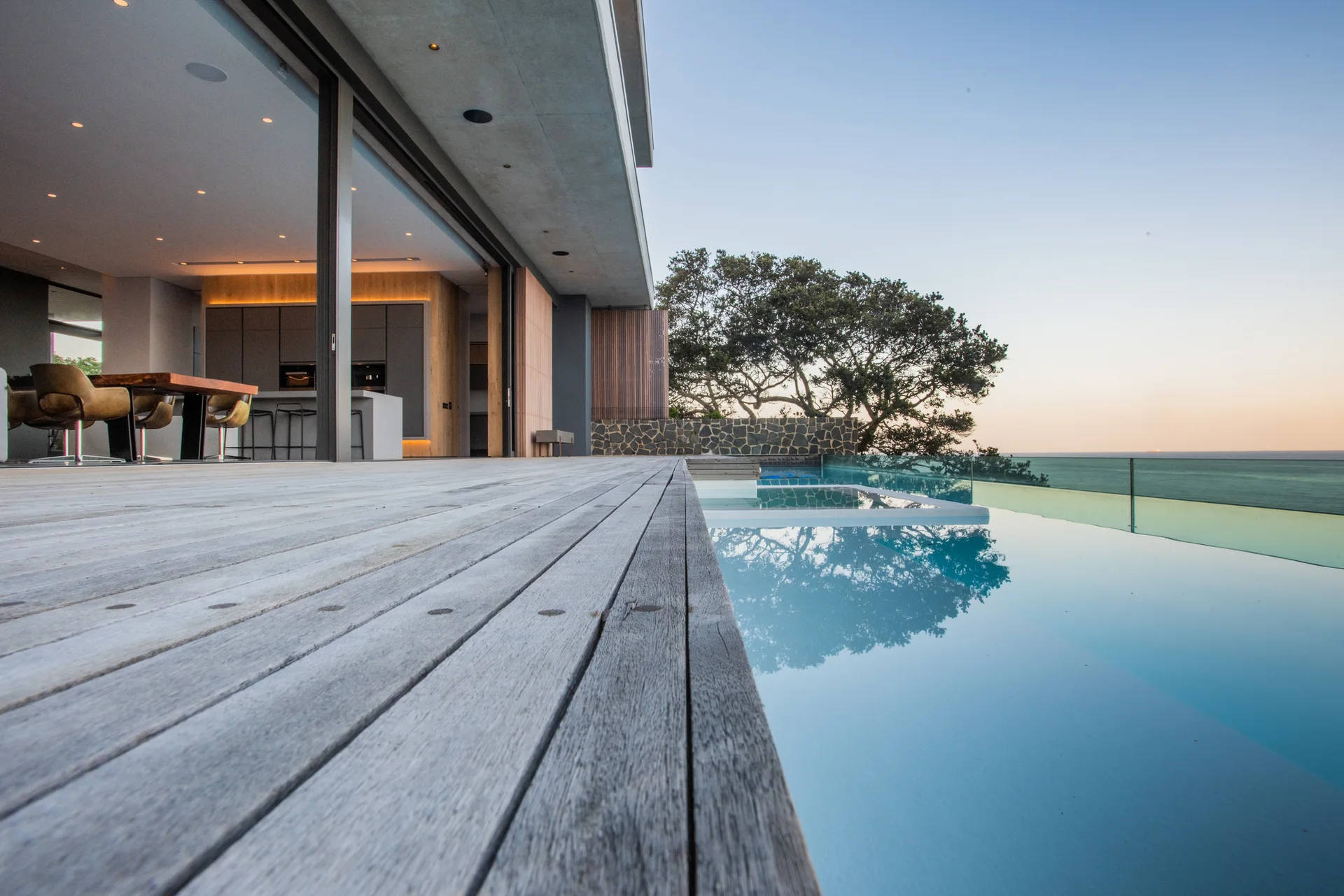 A weathered gray wood deck borders a blue infinity pool overlooking the ocean, with wide-open sliding doors leading to a modern interior.