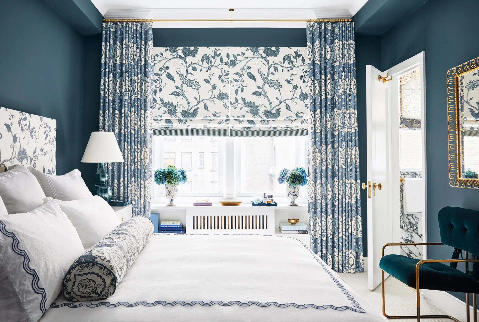 A blue bedroom features a bed with white scalloped bedding, coordinating floral curtains and shades, a built-in radiator cover, and a teal velvet chair.