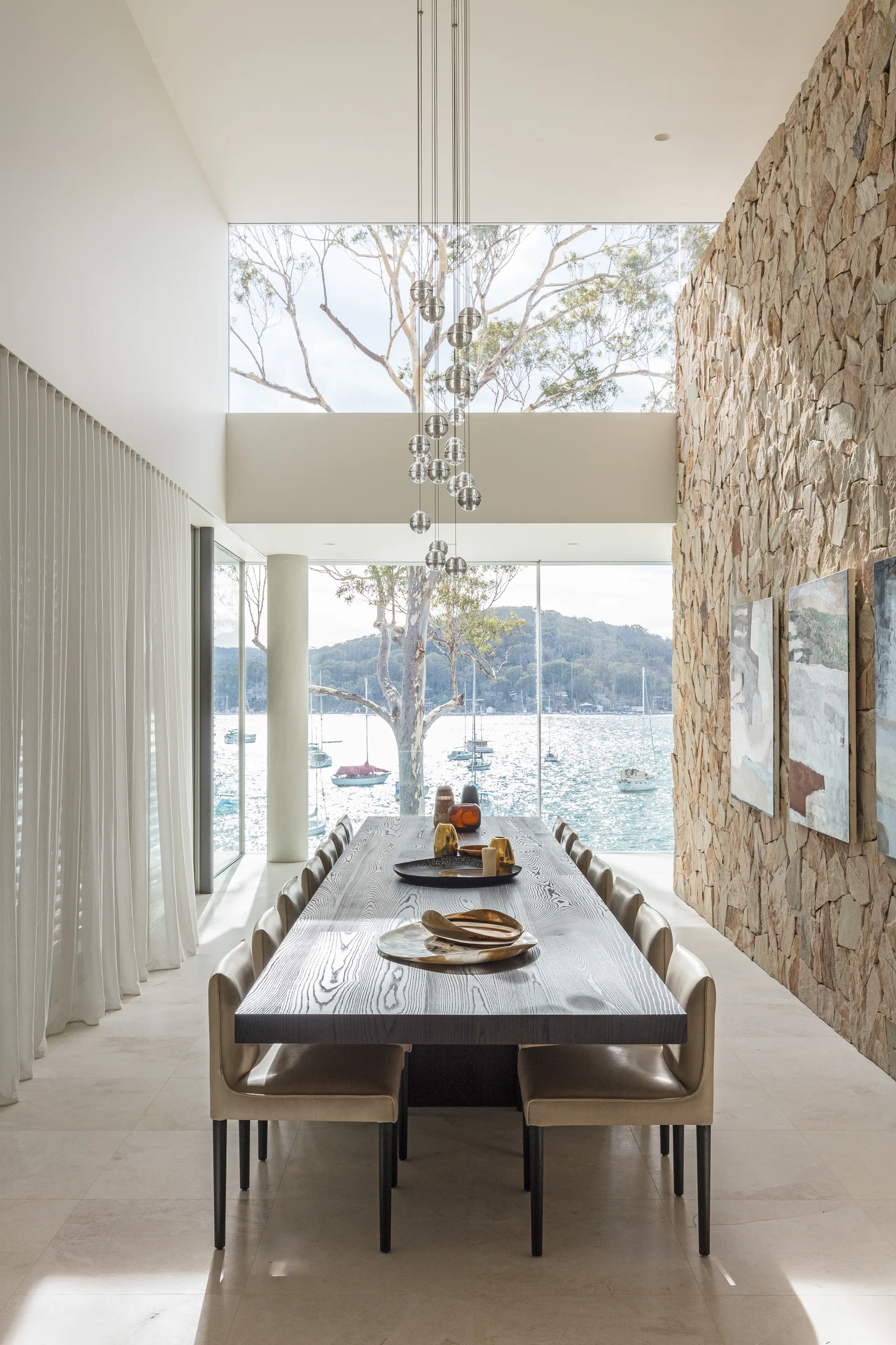 A modern waterfront dining room featuring a dark wood table with chairs next to a large stone accent wall and floor-to-ceiling windows.