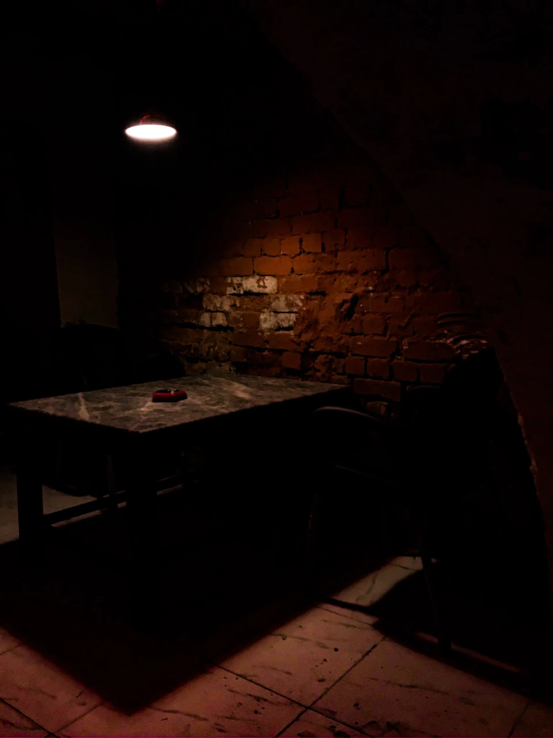A dimly lit interior corner featuring a stone table and chair set against an exposed red brick wall under a single glowing pendant light.