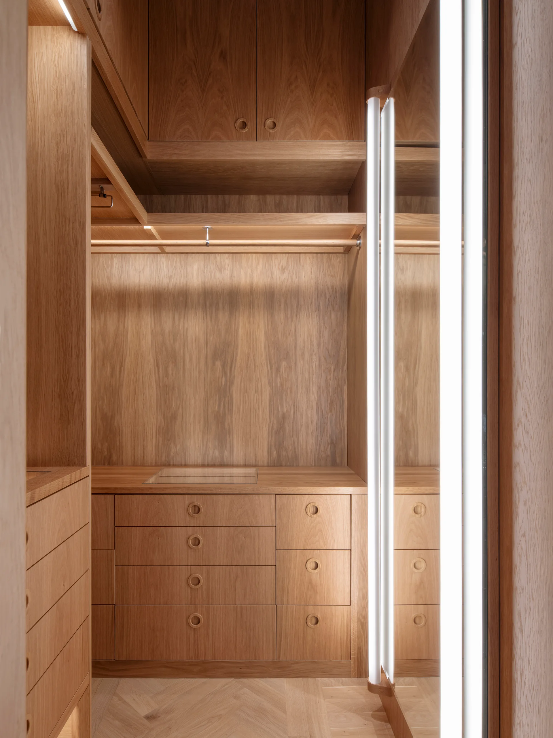 A custom warm wood closet with built-in drawers featuring circular cutouts, integrated vertical light strips, and herringbone flooring.