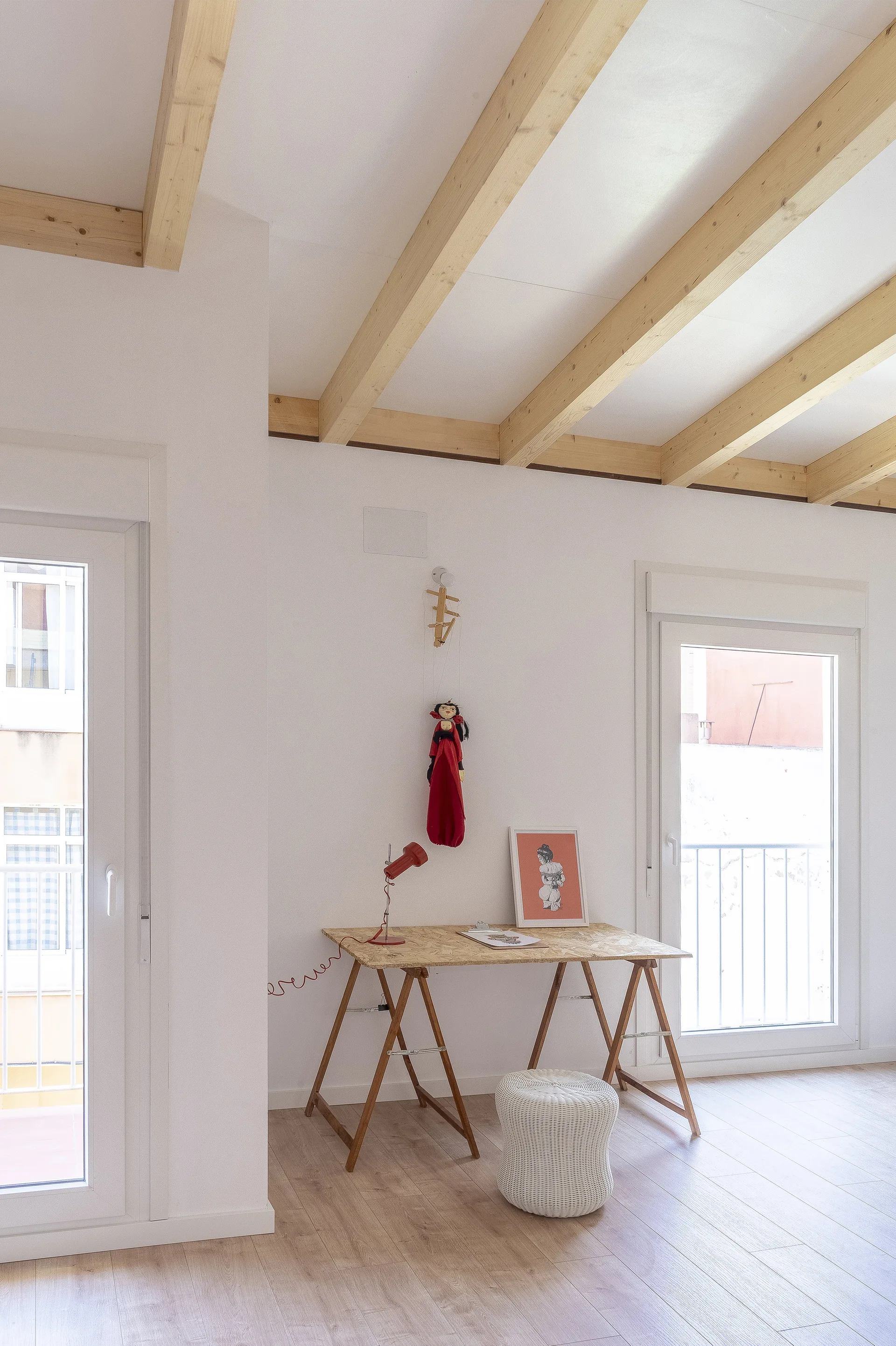 A bright home office features a light wood trestle desk with a red lamp, a white woven stool, and exposed ceiling beams between two glass doors.